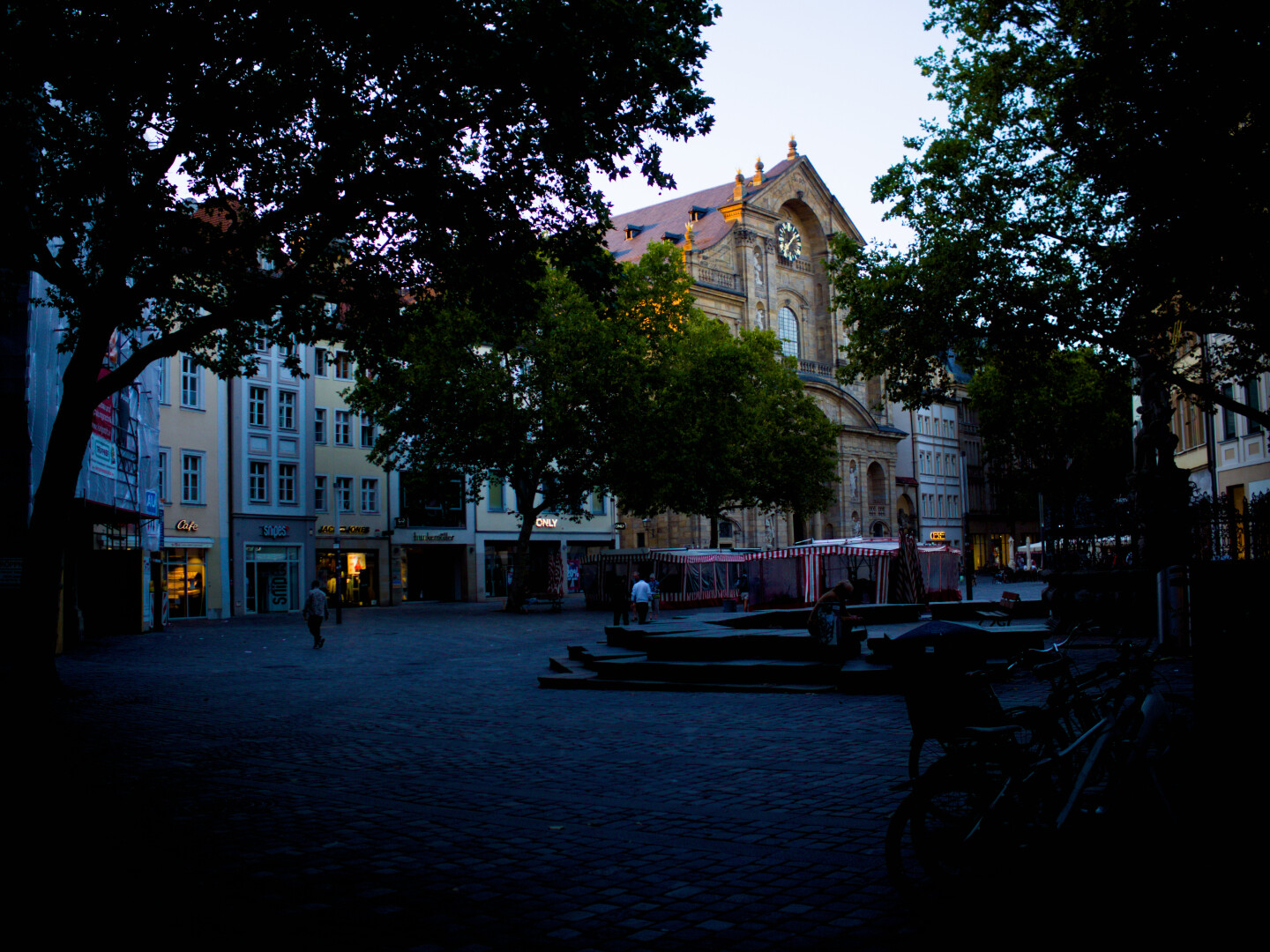 Fußgängerzone. Kirche geradeaus. Alte Häuser/Geschäfte. Bäume mit Blätern. Rechts eine Stizgelegenheit aus Stein. Markstände.