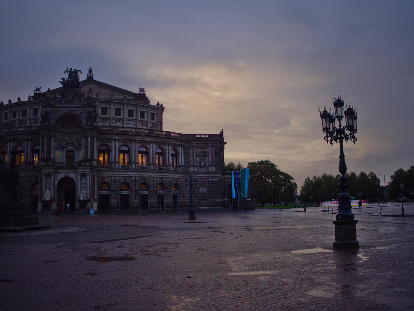 Die Oper seitlich Fotografiert. Rechts ein pompöser Leuchter. 2 Fahnen Mittig. Rechts in der Ferne Bäume. Bewölkung mit Strucktur. Plastersteine. Nass. Düster