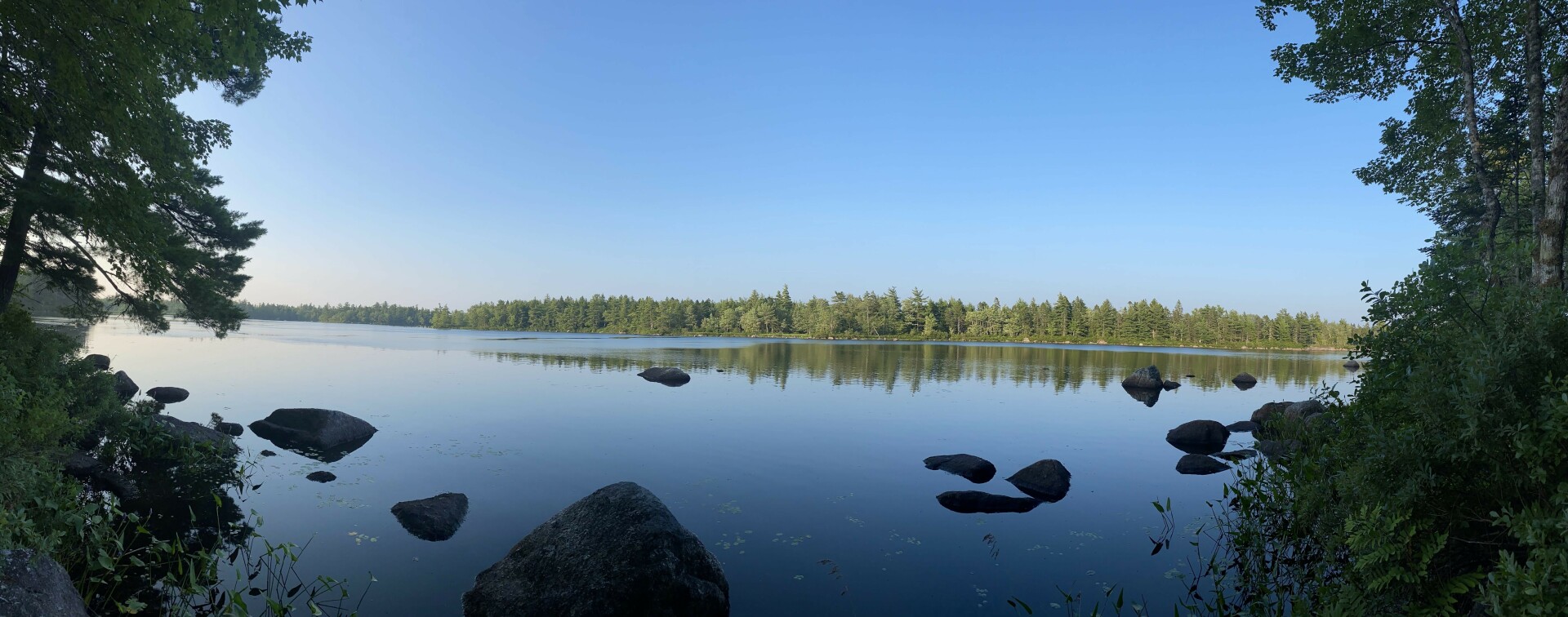 Still lake with rocks poking out from the lake. Trees either side and on the far shore. Trees on the far shore reflected in the water. Blue sky above the lake and no clouds.