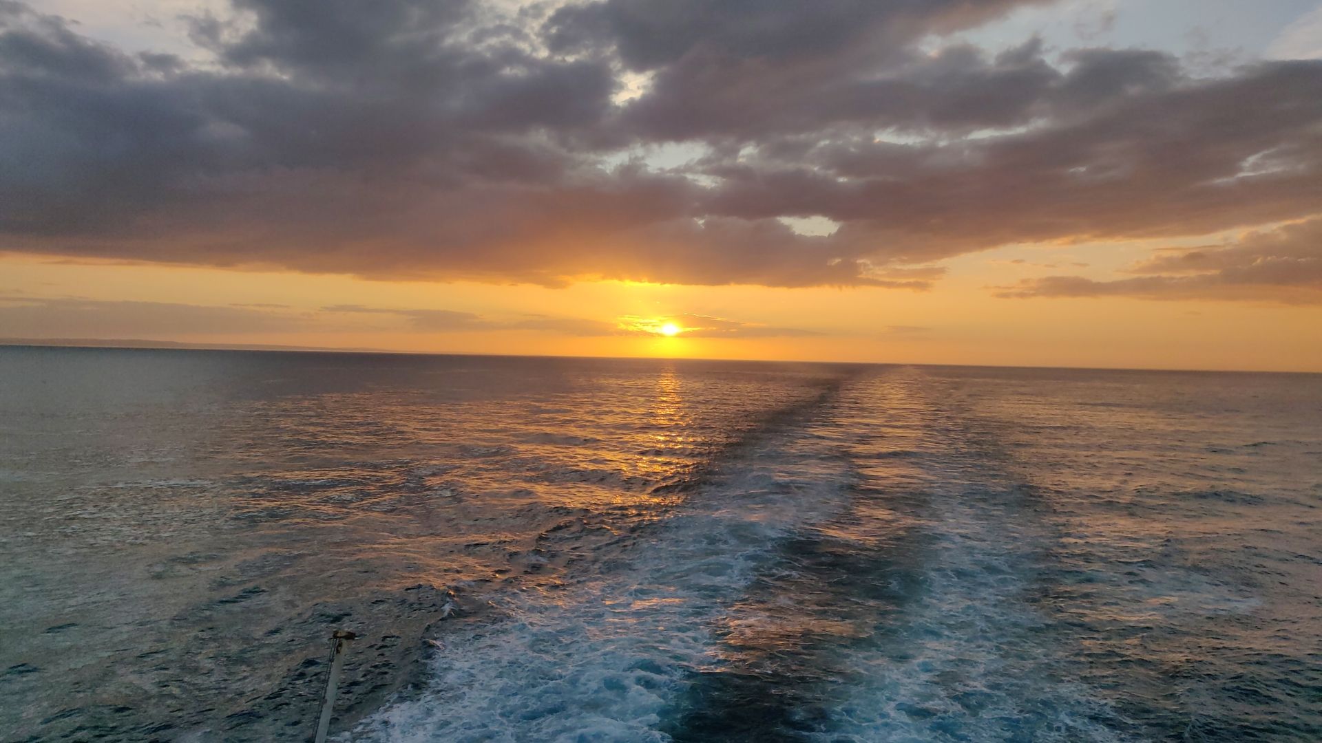 Photo of the ocean at sunset. The sea is a deep blue with white frothy parallel streaks in it from a boat that go into the distance. The clouds are dark and covering around 60% of the sky in clumps. The sun is a small orange dot on the horizon, turning the sky around it a yellowish orange