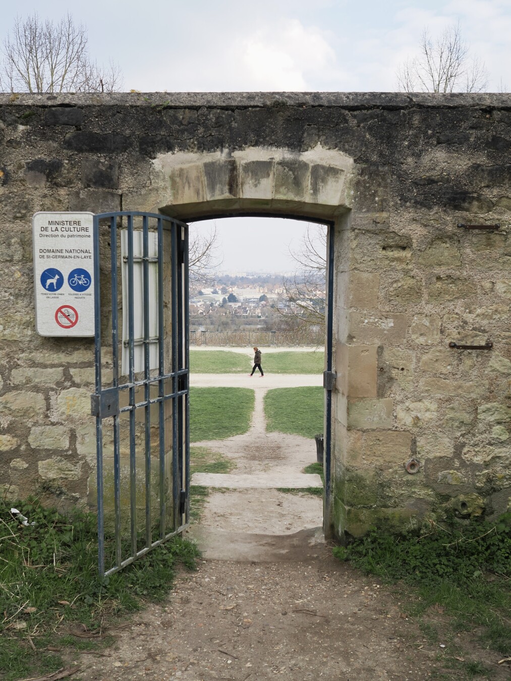 A photo of a passage in an old stone wall with an open metallic grated door. There is a dirt crossroad visible in the passage, with a single person walking from right to left. Further beyond, there is a drop and many houses going until the horizon covered in slight haze or smog.

On the wall, there is a sign with the following text: Ministere de la culture.
Direction du patrimoine.
Domaine national de St-Germain-en-Laye.

There are three icons visible: two that allow dogs (but with leashes) and bicycles (with reduced speed), and one that disallows picnics.