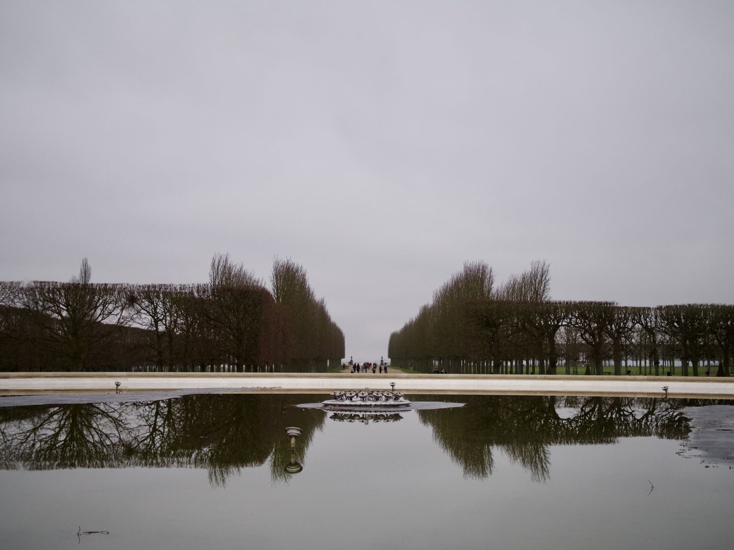 A photo of a park, view is over a fountain surface (not working), with a path with trees on both sides behind it. There are people on the path far in the distance, maybe around 20 of them, with the path ending almost at the horizon, where a grey sky takes over and fills the top half of the image. It is a twilight, the photo is mostly grey, with a bit of green grass visible in-between the trees. The fountain surface takes the bottom quarter of the photo and reflects the tree and the sky.