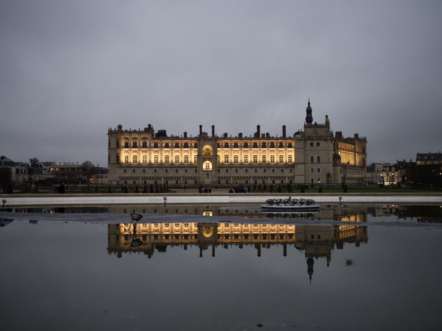 A photo of a chateau with its reflection in the surface of a fountain. The building is highlighted by yellow lights which outline a row of windows. A bit of city houses are visible on both sides of the chateau.