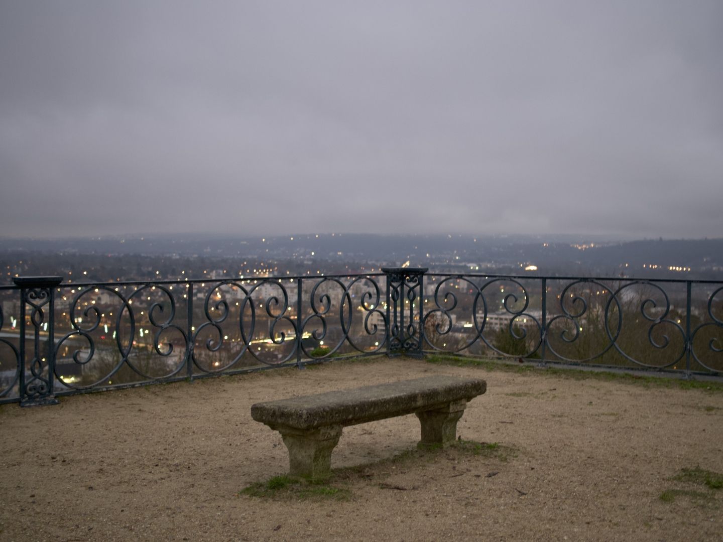 A photo of a bench standing in sand on a corner of a high terrace overlooking a city below. It is twilight, city is full with blurry yellow lights, a bit of a river with a bridge can be visible below as well. Between the city and the bench there is a metal fence with a swirly pattern reminiscent of butterflies. A hilly horizon is covered by grey clouds.