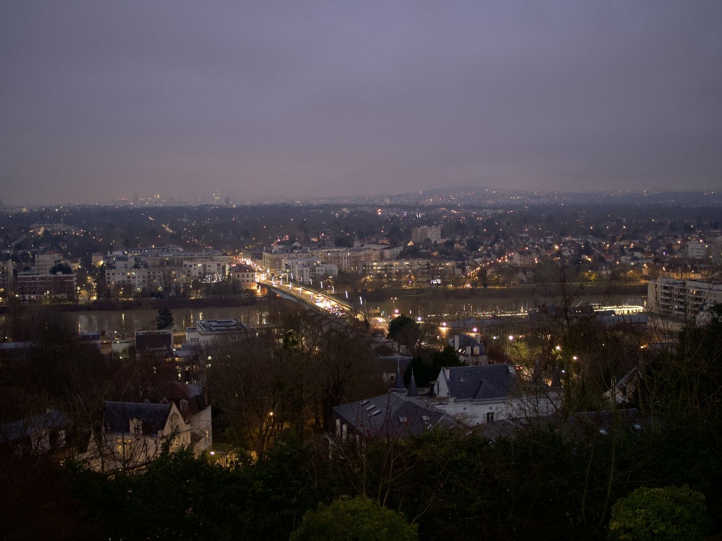 A photo of a view from the terrace over the river, a bridge over it, and the city before and further back. It is darker than on the previous photos, and more yellow lights are visible, and they are now in focus. The front of the photo is darker, with roofs of closer buildings visible from behind leafless trees. The sky is grey, there are hills on the horizon merging into the greenness of the night and littered with buildings.