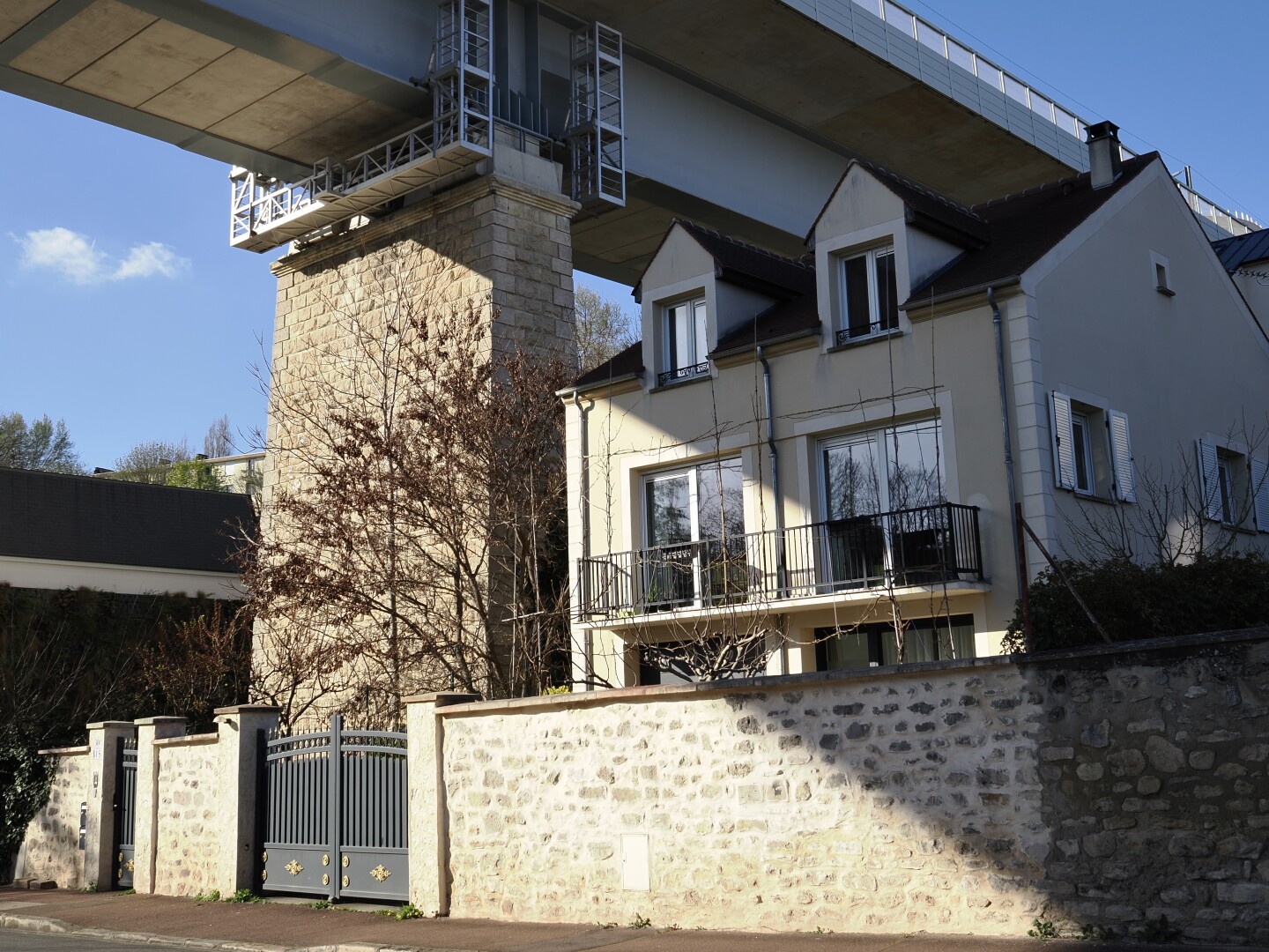 A photo of a two-storey house under a railway bridge. The sun is shining brightly, with just a single cloud in the blue sky, but only a part of the house is light-up by it, with a shadow of a bridge going diagonally over the building and a stone fence near it.