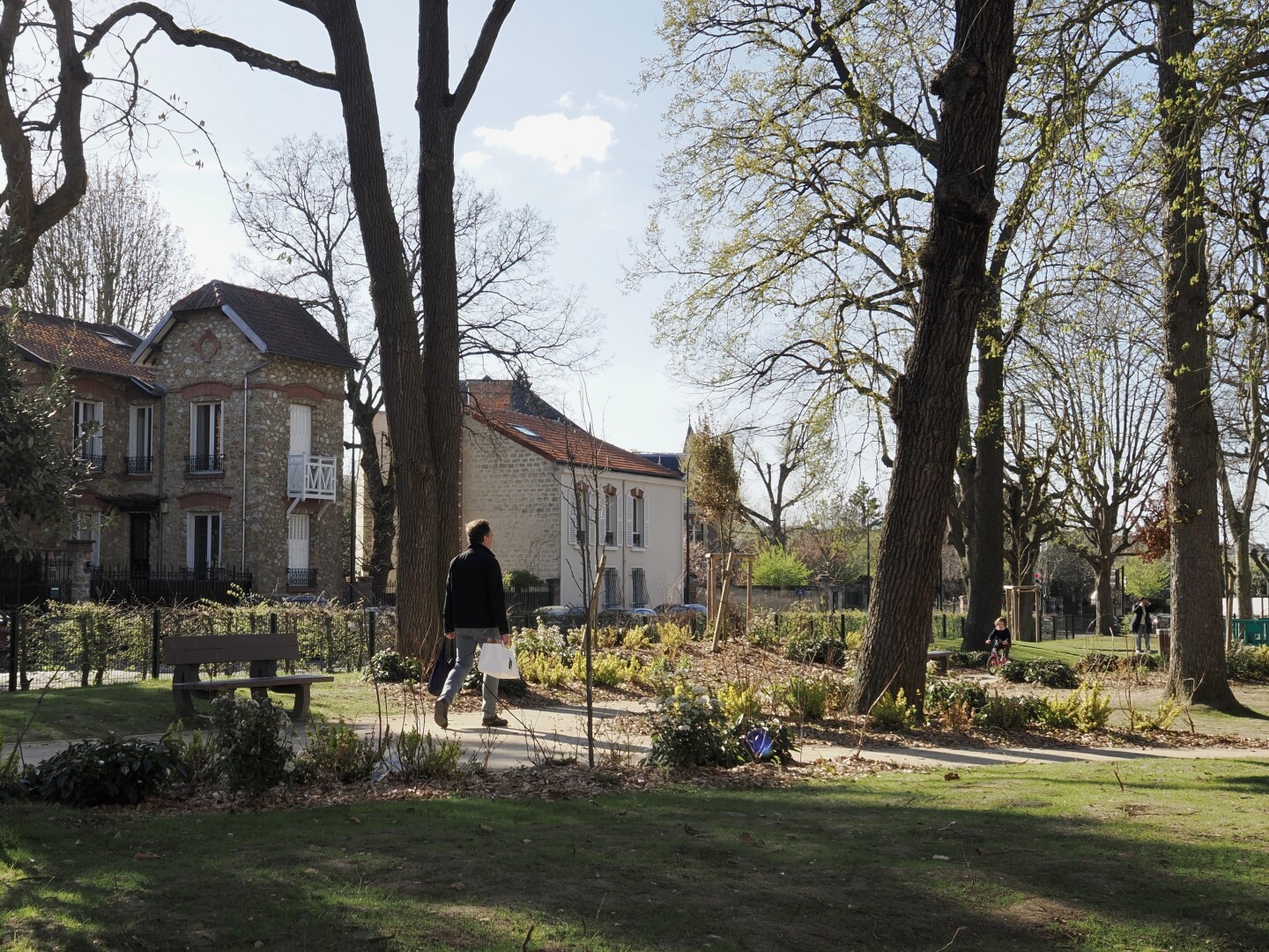 A photo of a small park with some houses visible nearby. Close to the middle, a person is walking to the right, holding a paper bag in one hand, and a clothe bag in another. Further away on the path, there is a little girl on a pink bicycle. Even further there is a playground and another person can be barely seen there. There is a lot of green in the photo: grass and leaves, and the sun is shining brightly, leaving many shadows. The sky is mostly clear, with a single cloud barely visible.