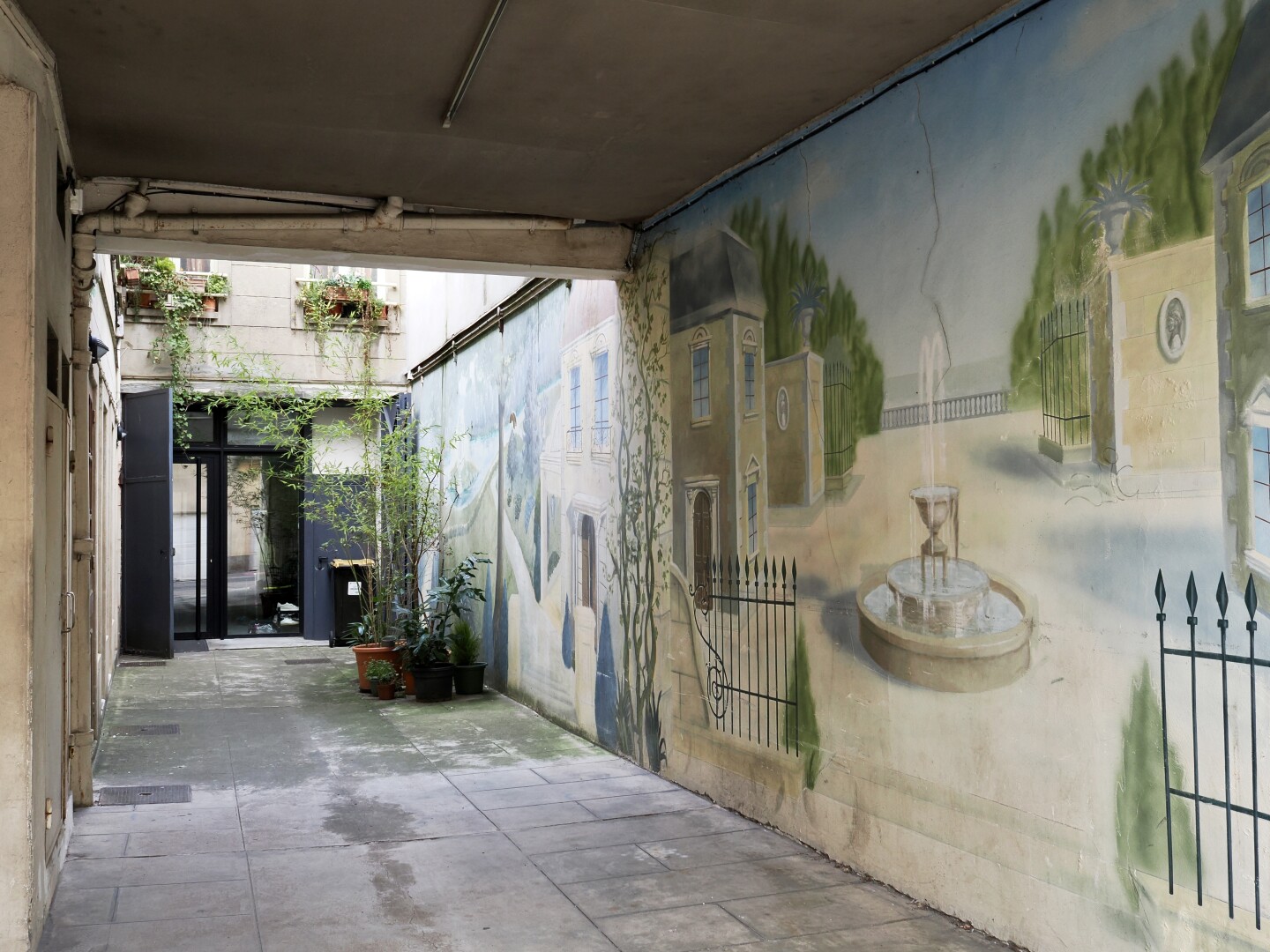 A photo of a small courtyard, with graffiti or paintings. One shows a courtyard near a terrace with a fountain in the middle, another shows a park near a lake or a river. Further in the courtyard there are a few pots with plants on the windows higher up, and on the floor, with green vines connecting them.