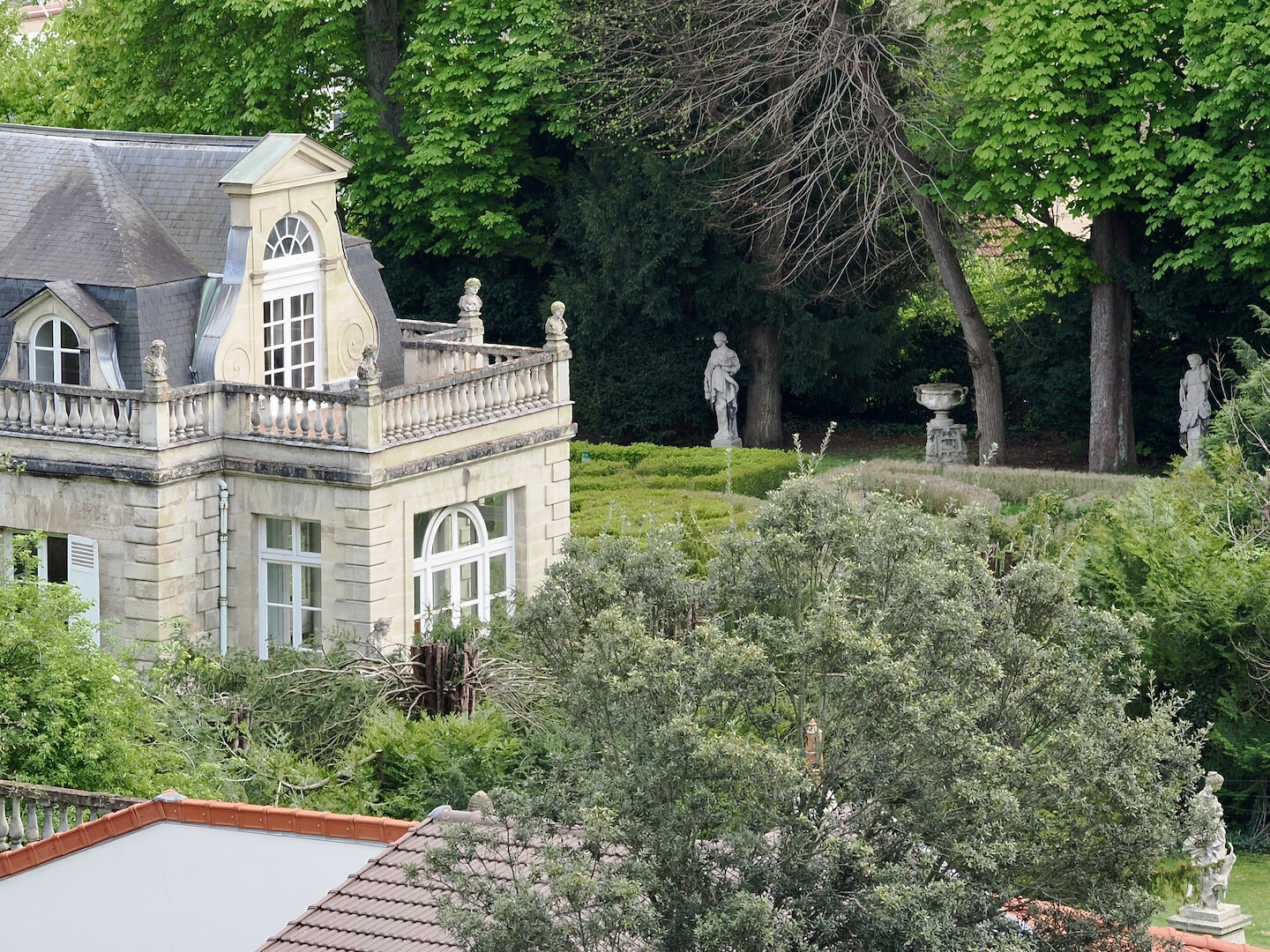 A photo of a courtyard of an old building shot from a bit higher up. The frame is surrounded by trees with green leaves. A hedge labyrinth visible behind the building. There are various statues: three visible in the garden, and four busts visible on the balcony.