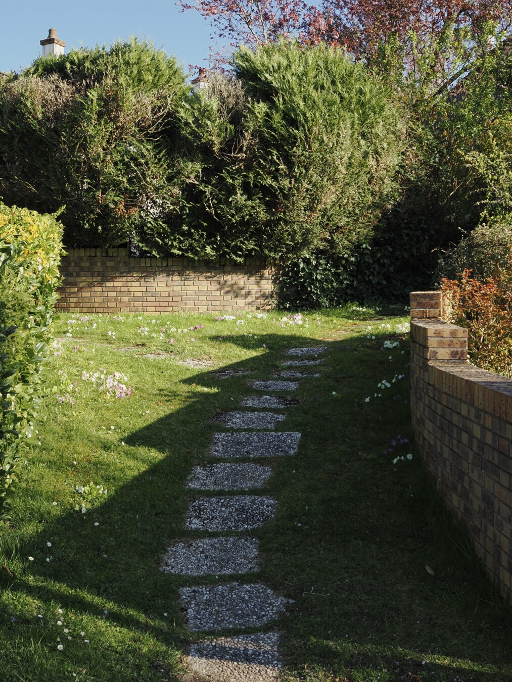 A photo of a path, going in a slight curve a bit uphill. The path consists of large white stone squares visible from between the grass. On the left there is a hedge, on the right and forwards there are brick fences. Behind the forward fence there are trees and bushes, with two small chimneys visible. The sun is shining brightly, the shadow of a stone fence on the right covers most of the path and creates angled ridges from its slight change in elevation.
