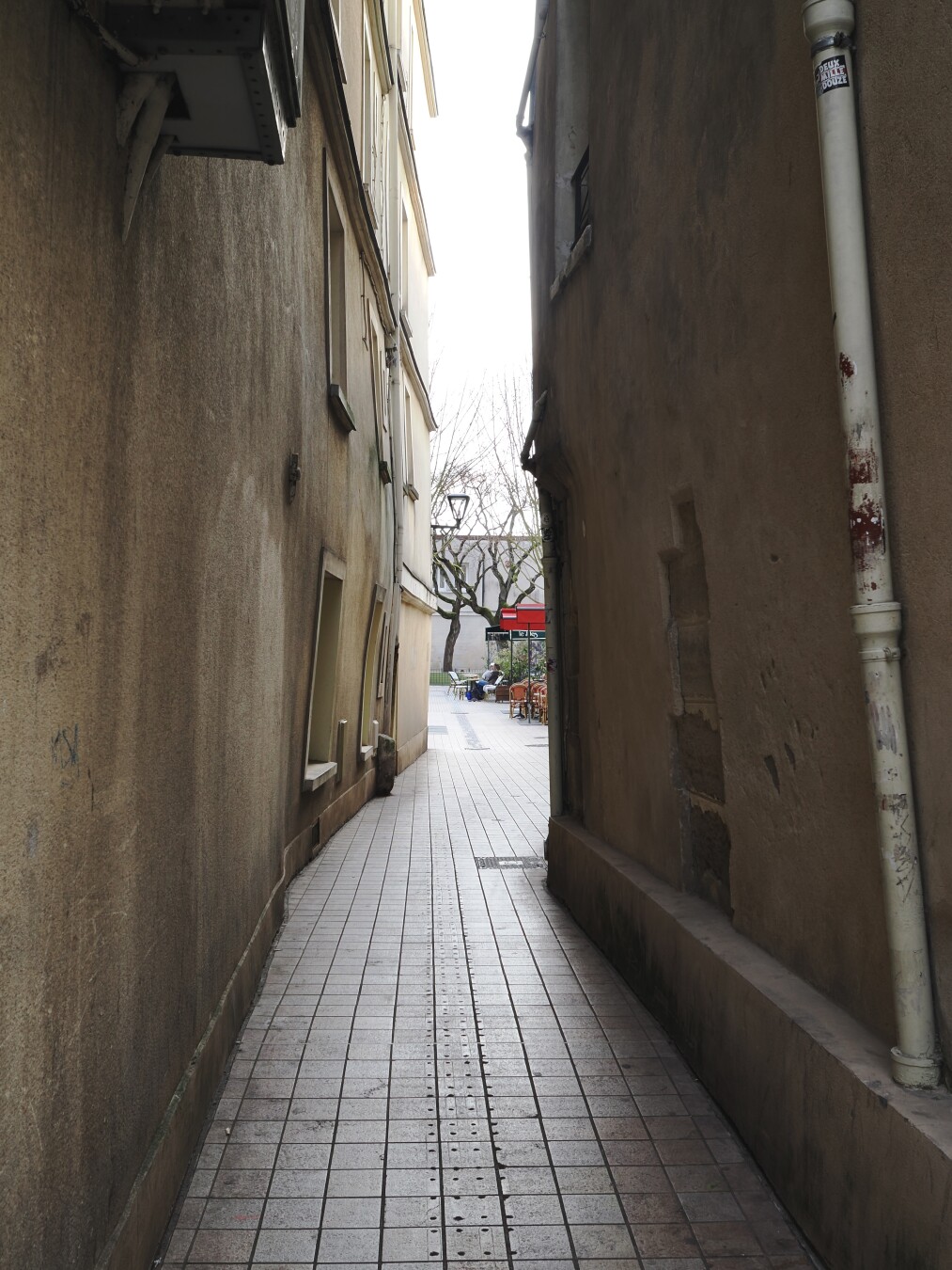 A photo of a narrow path between two old buildings, a bit warped with time. The floor is all tiled, the walls are plastered with spotty yellow paint. The path opens up to a small court, with something like a café: tables, awnings, chairs, and two people visible sitting. A bit further — some trees and a street lamp.