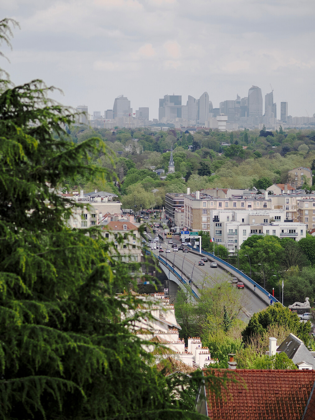 A photo of a view from a hill, with a bridge  going from the bottom right corner towards the center of the frame, followed by various buildings, then many trees with a cone roof peeking through them, and skyscrapers on the horizon. The photo is framed by an out-of-focus fir on the left, and there is a red tiled roof visible in the bottom right. The sky is grey with clouds.