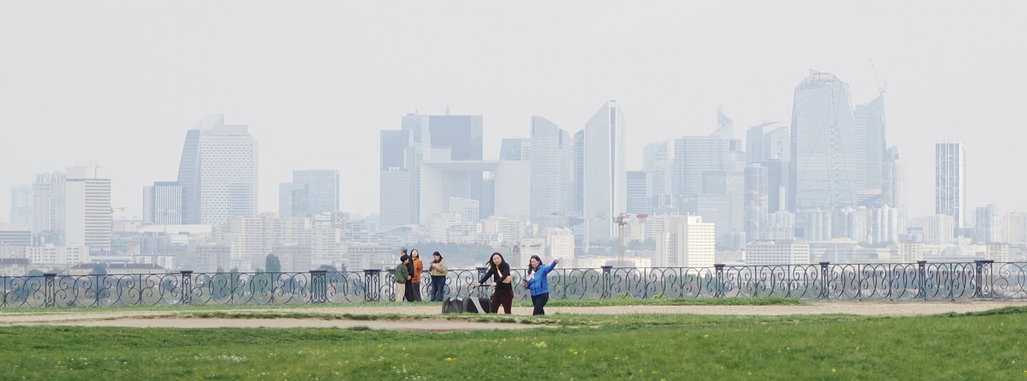A photo of a few people walking on a terrace of a park, with a background full of skyscrapers of La Défense in the distance. The photo is made with a telephoto lens, resulting in the skyscrapers being seemingly closer to the viewer than they are in comparison to the people. Closer to the camera, there are two persons, one is rolling a wheelchair, another is pointing at something. Further, near the transparent metallic fence, there are four more people. The sky is gray, with the skyscrapers nearly merging with it.