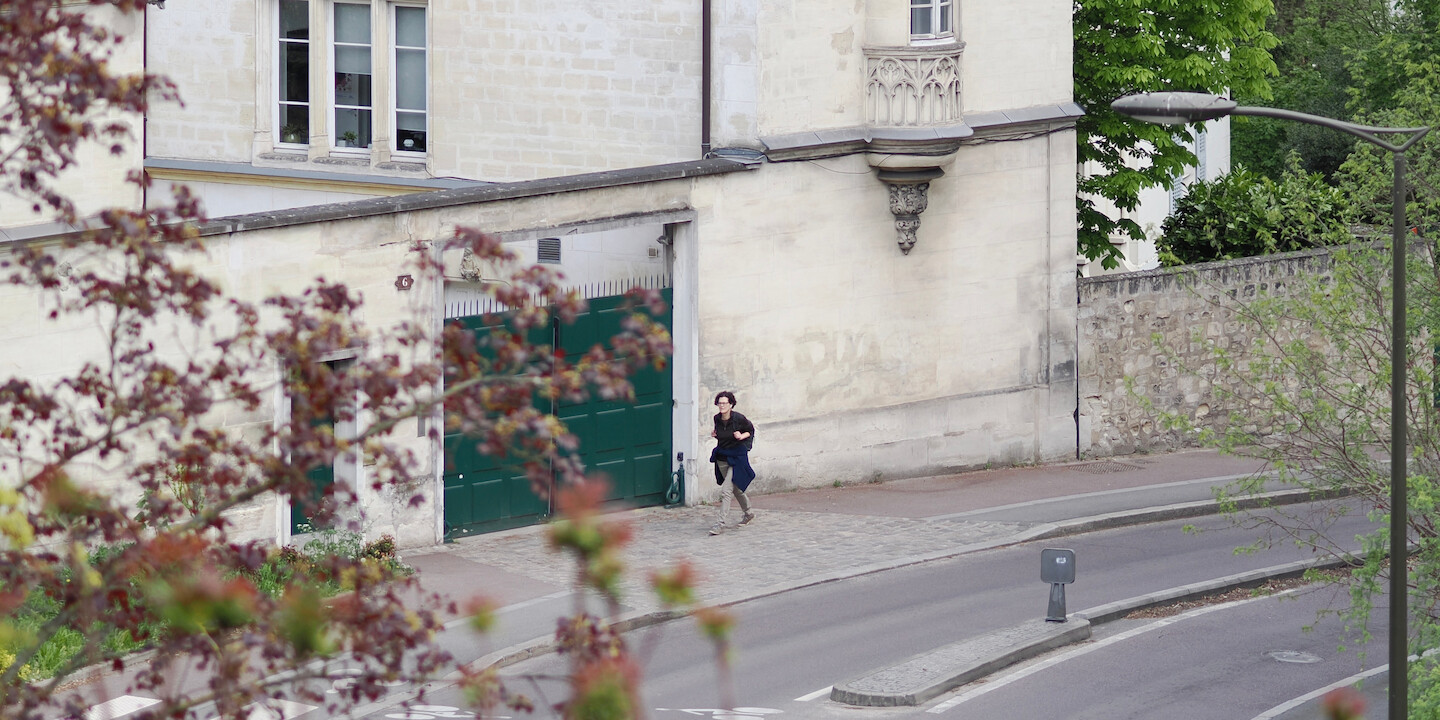 A photo of a person walking a bit uphill near a road. Behind them, an old brick building, painted white, with a courtyard separated by a wall with two green doors: for people, and for cars. On a side of that building, there is a small round oriel window with ornamental decorations under it, and faded graffiti below. On the two-lane road, there is a small safety island in the middle with a back of some sign. The right of the photo is framed by a streetlamp and some green leaves of trees. The left of the photo is covered by red, with a bit of green out-of-focus leaves.