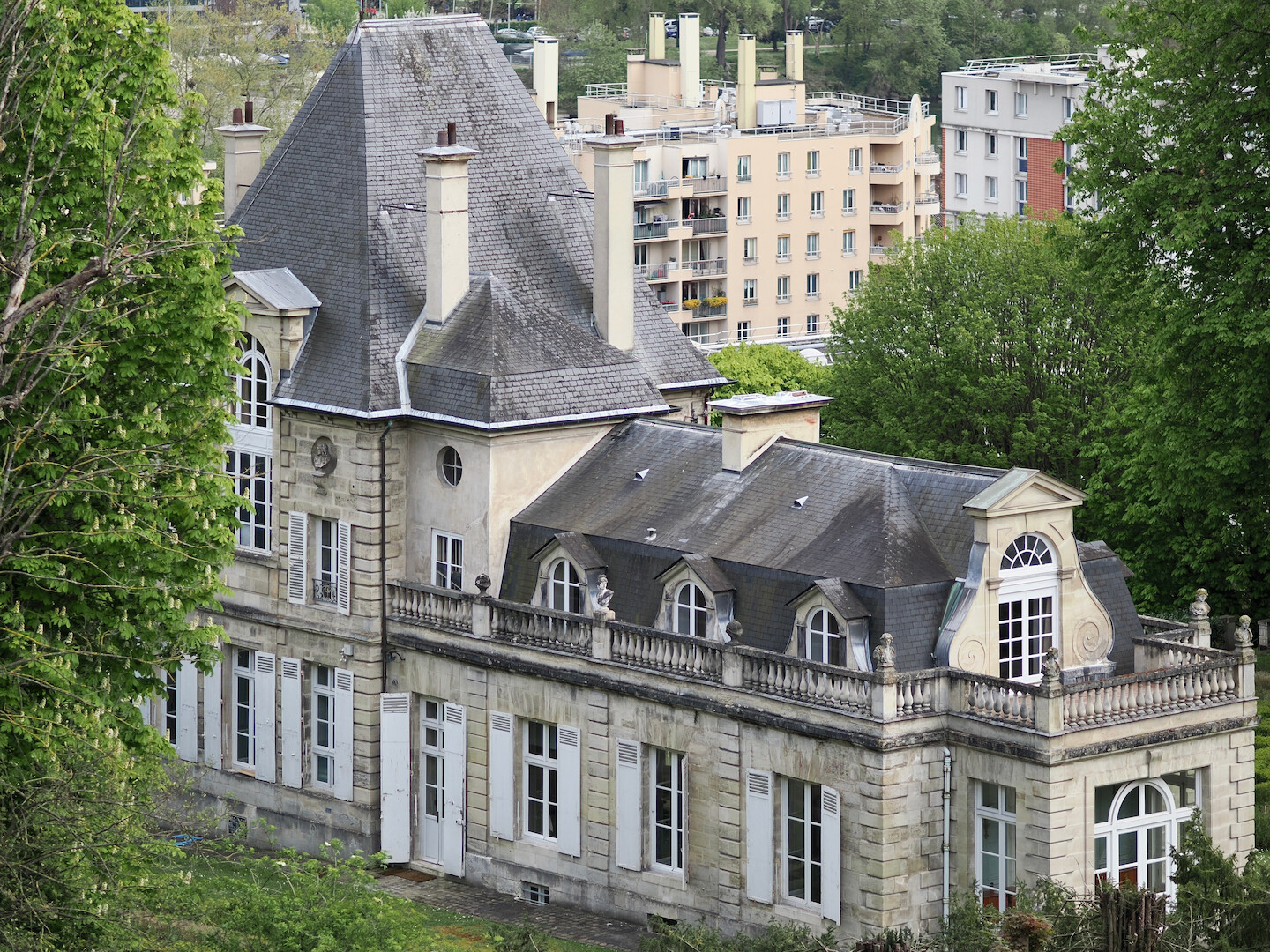 A photo of “Pavillon Sully” — an old building with steep grey tiled roofs, chimneys, and some statues visible on top of its walls. It is surrounded by the trees, and with some more modern buildings visible in the background.