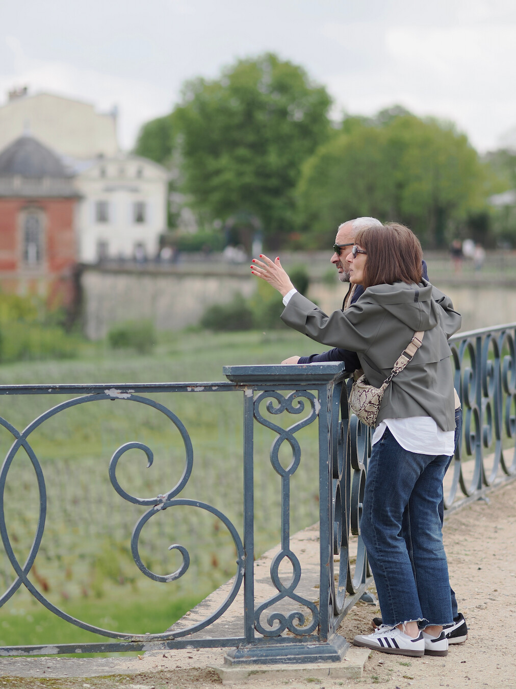 A photo of two persons standing near a fence of a terrace, looking into the distance. One of them is gesticulating with her hand, as if pointing towards something. The pair is in focus, as well as the dark green painted and slightly dilapidated metal fence with swashes. The rest is out of focus, with an old building visible on the left, and a few trees visible on the right. The sky is overcast and is bright grey.