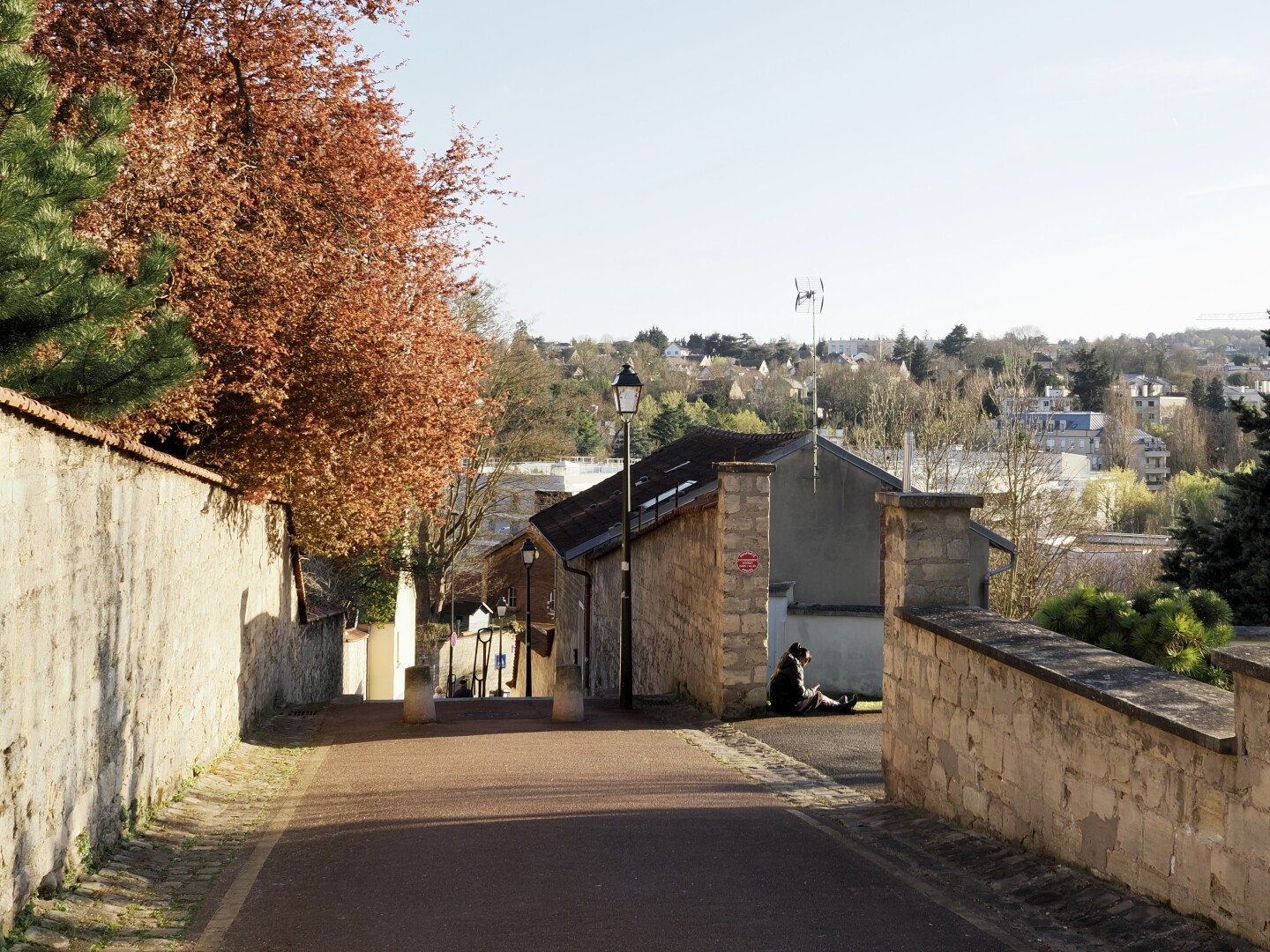 A photo of a stair path from a steep incline, with many houses between the trees visible down below on the horizon in the background. The sun is soon to be set, and it lights up brightly the scene. There are walls on both sides of the path, with an opening on the right. A woman is sitting with her phone in the opening. Trees are visible on the left, behind the wall —&nbsp;a few green branches of a pine or a spruce, and orange leaves of some other trees. Some other small details in the shot: a low streetlamp near the stairs, a couple of people barely visible descending them, some signs, a house near a way visible through the opening, and an antenna on top of its roof.