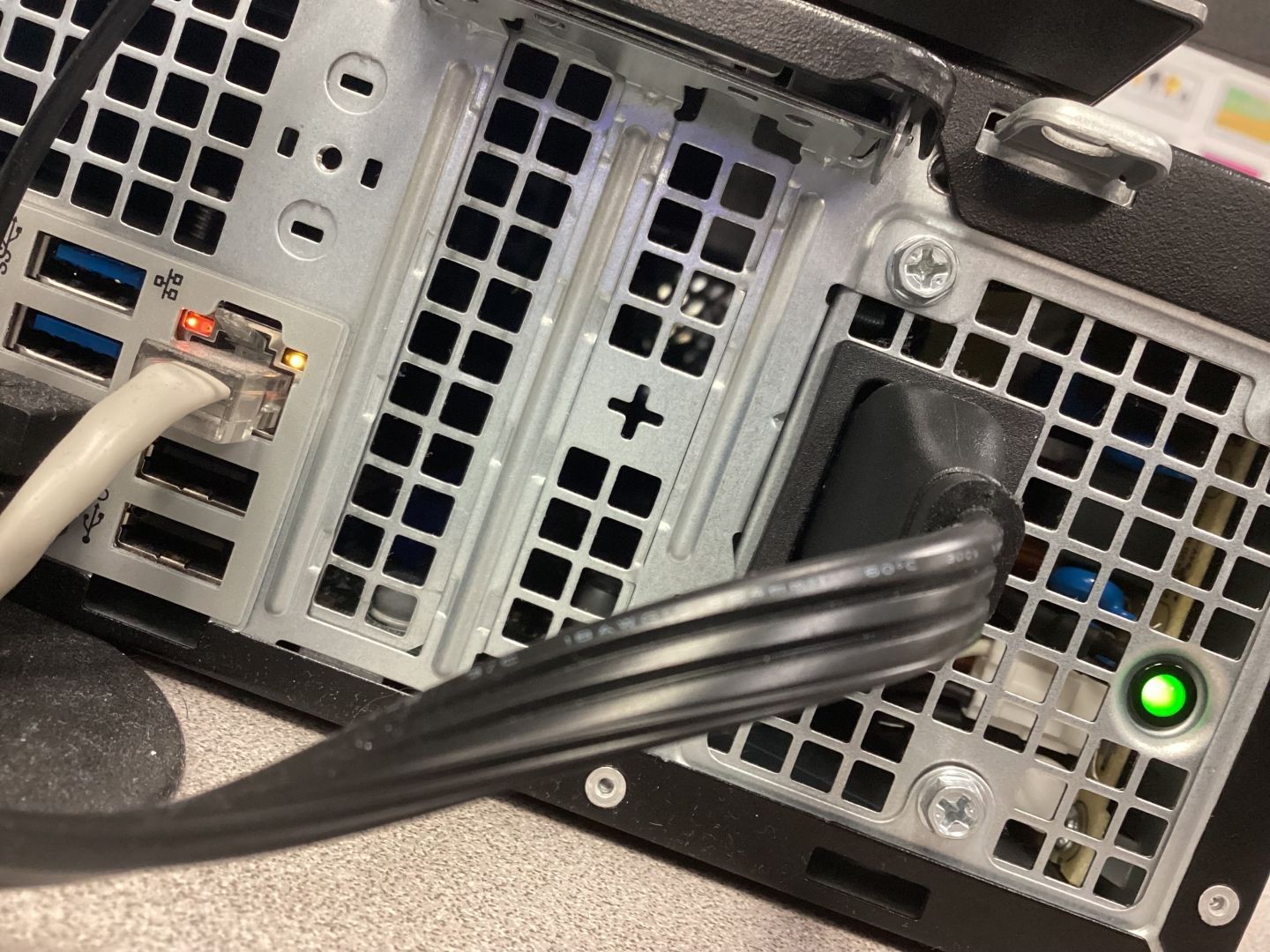 An image of the back of a modern Dell Optiplex at a relatively extreme dutch angle.
The Ethernet port is glowing, and a light on the power supply is on.
The back is grey metal with a grid pattern of squares cut out for airflow.