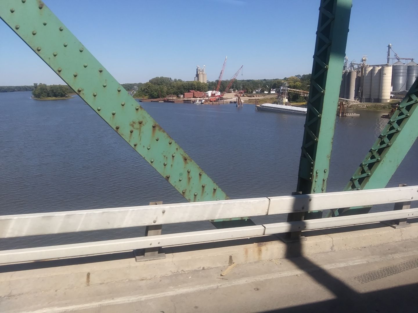 A green painted metal bridge over a river framing a distant industrial plant of some sort.
