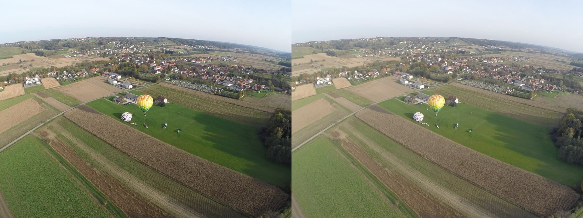 View from a hot-air balloon about 100m above ground. There is a village in the middle distance and a ridgeline behind it. On the field below, one other balloon is about to depart, and a third is still on the ground ready for inflation.