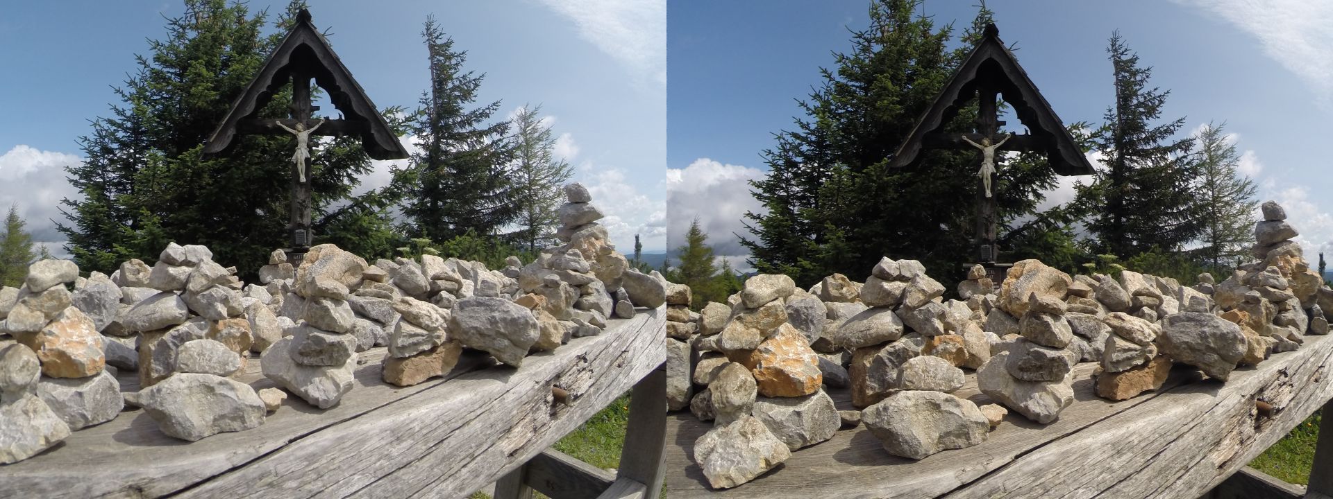 A wooden table full of stacked rocks near a wayside cross by a hiking trail. There are at least 30 such stacks, neatly arranged in rows. The cross is flanked by some trees; the weather is sunny with scattered clouds.