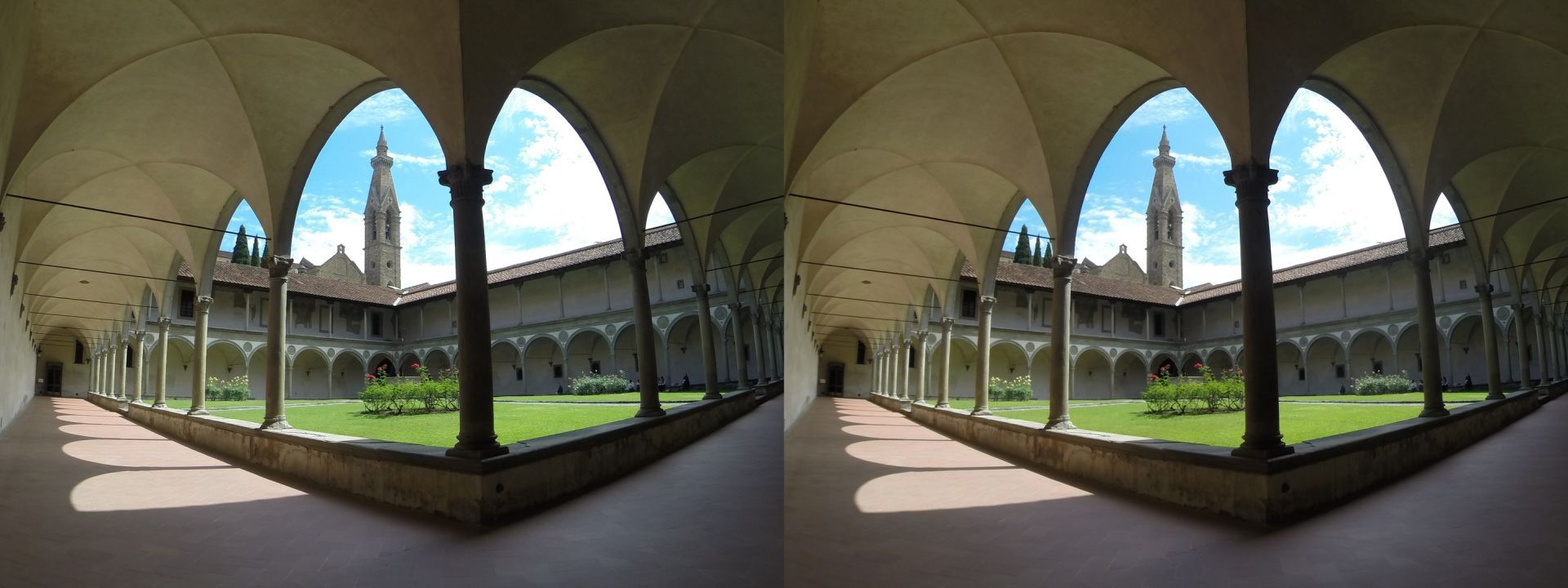 Stereoscopic image of a square courtyard on a sunny morning. It is bordered on all sides by arcades, in its centre is a patch of green grass criss-crossed by a walkway and dotted with rose bushes. In the background, the belfry of the Santa Croce basilica is set against a blue sky with scattered white clouds.