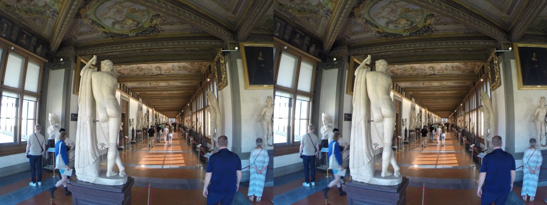 Cross-view stereoscopic image of the view down a long and wide corridor on the second floor of the Uffizi gallery. A nude marble statue has its back toward the camera, as do several (clothed) visitors. The corridor is illuminated by sunlight flooding through the large windows. A row of statues each is on either side, with explanatory signage beside them. The ceiling is decorated with elaborate coffering.