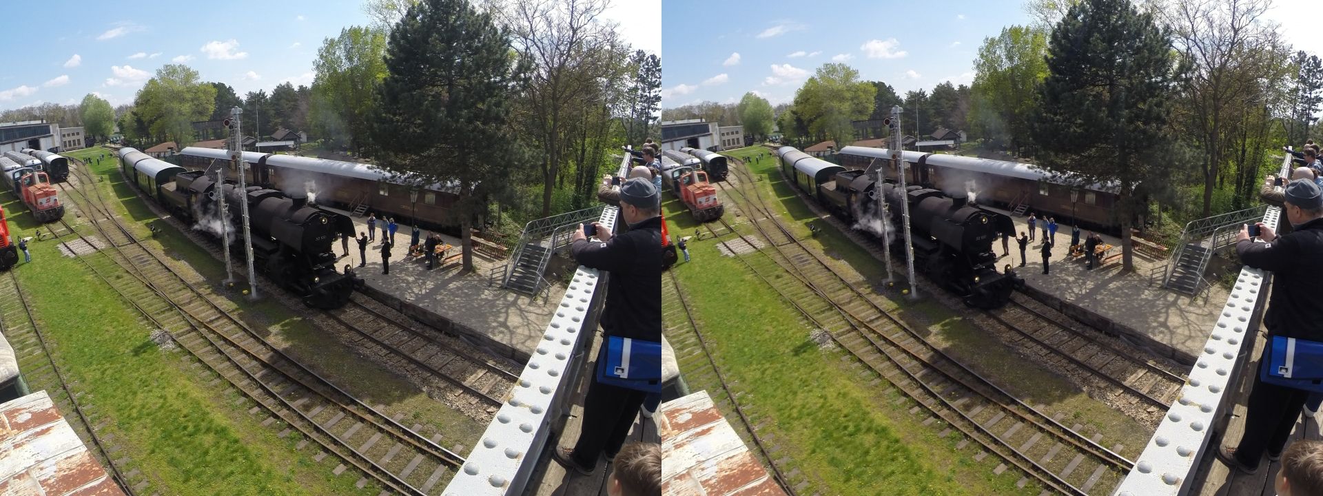 A steam locomotive pulling three passenger cars, crossing the shot from top left to bottom right, viewed from an footbridge above the track on a bright, sunny spring day. Some onlookers stand on a platform below while the train passes, and other photographers are snapping pictures with their mobile phones and cameras from the footbridge.