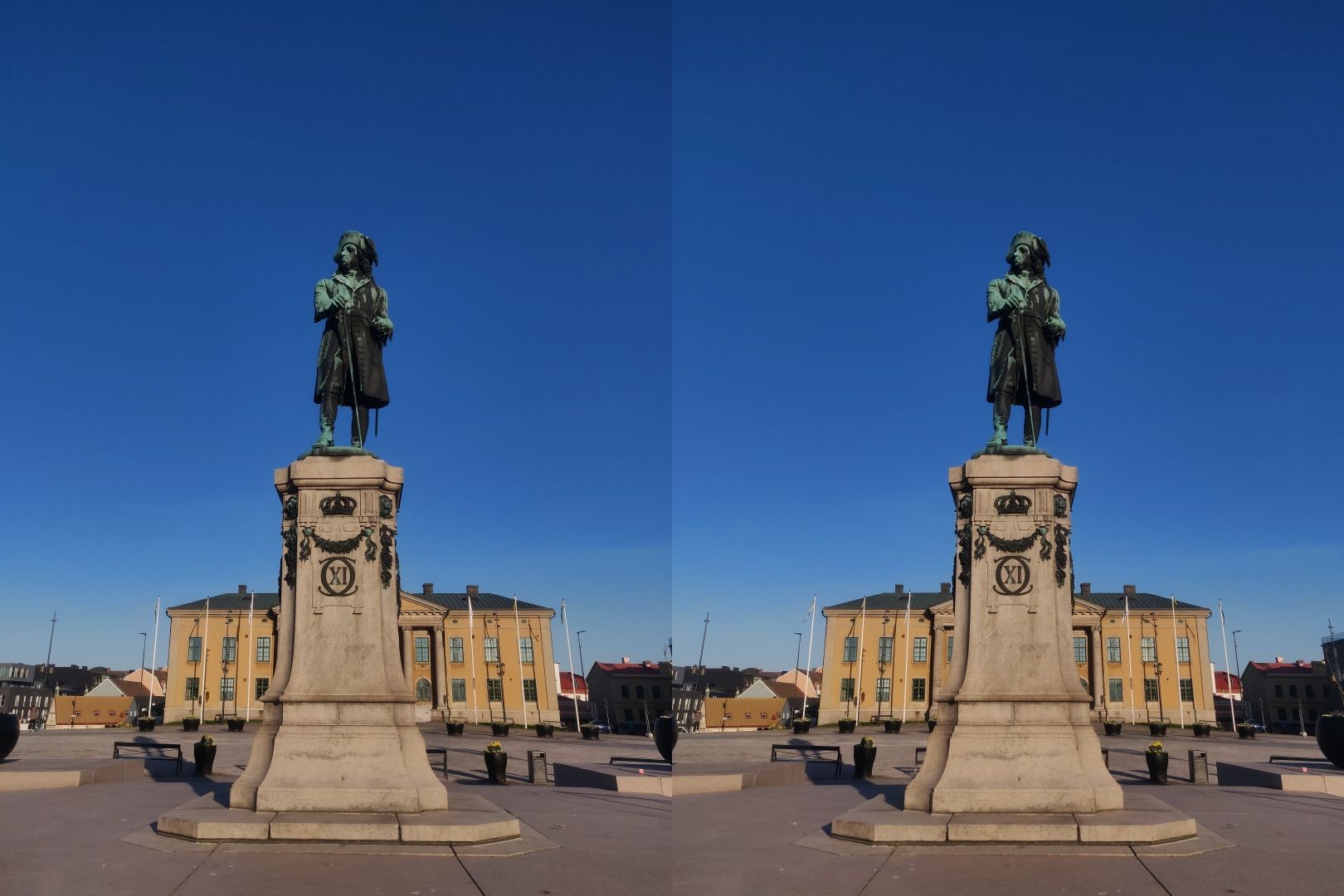 Cross-view stereoscopic image of the monument to Sweden's king Charles XI in Grand Square (Stortorget) on the island of Trossö, in the Swedish city of Karlskrona. The sun is low in the east on an early spring morning, and the sky is clear with a deep shade of blue. Behind the statue there is a row of flagpoles, and the building housing the Blekinge district court (Blekinge tingsrätt). The king's statue on the pedestal depicts 17th century court attire, and is made of bronze. The roman numeral XI is on the front of the pedestal.