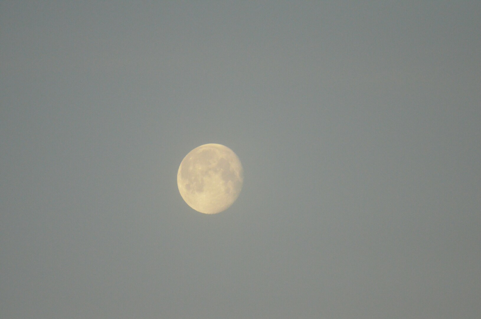 Picture of the moon with dark craters surrounded by a sky which has a pale grey hue.