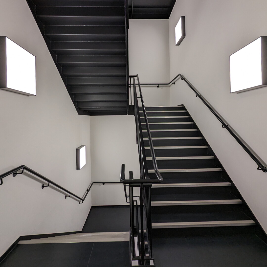 A photo of stairs at an office building going down on the left and up on the right. The area is lit with rectangular lamps. The stairs are black while the walls are white.