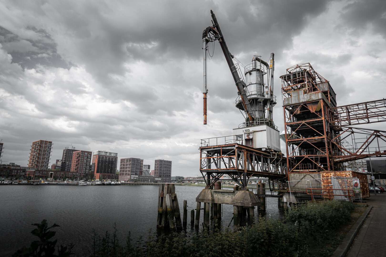 Old rusty crane on a wharf. Right a road, left the water of the harbour. Some tall buildings on the other side of the harbour.