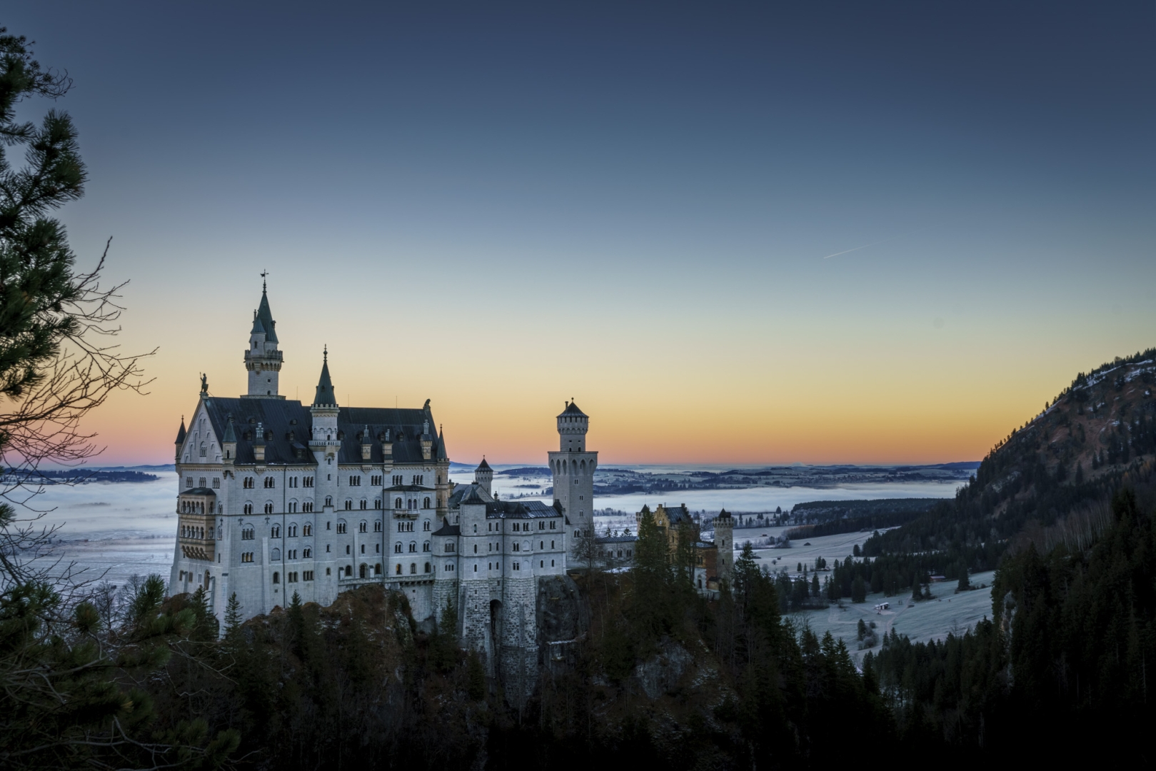 White castle on a rock in front of fields covered with fog. Orange blue sky above it all