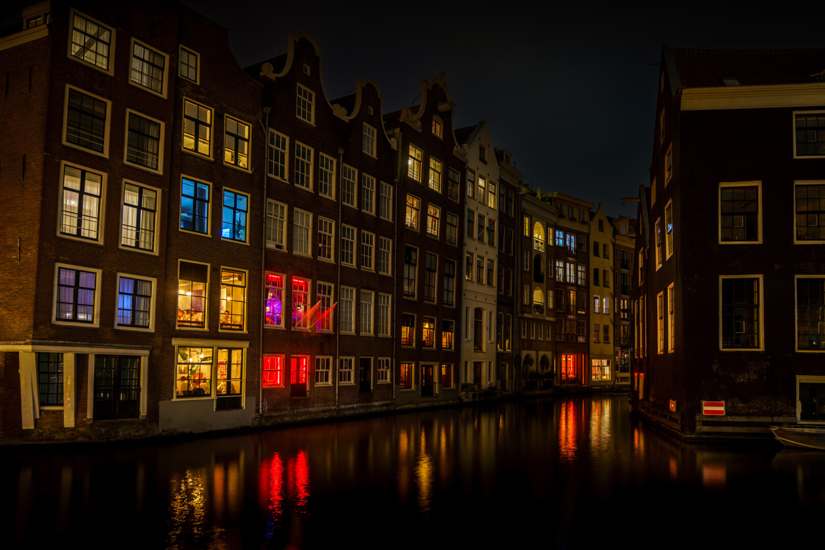 Old tall houses around a junction of canals. It is dark, and from the windows you see different coloured lights. The scene is reflected in the water.