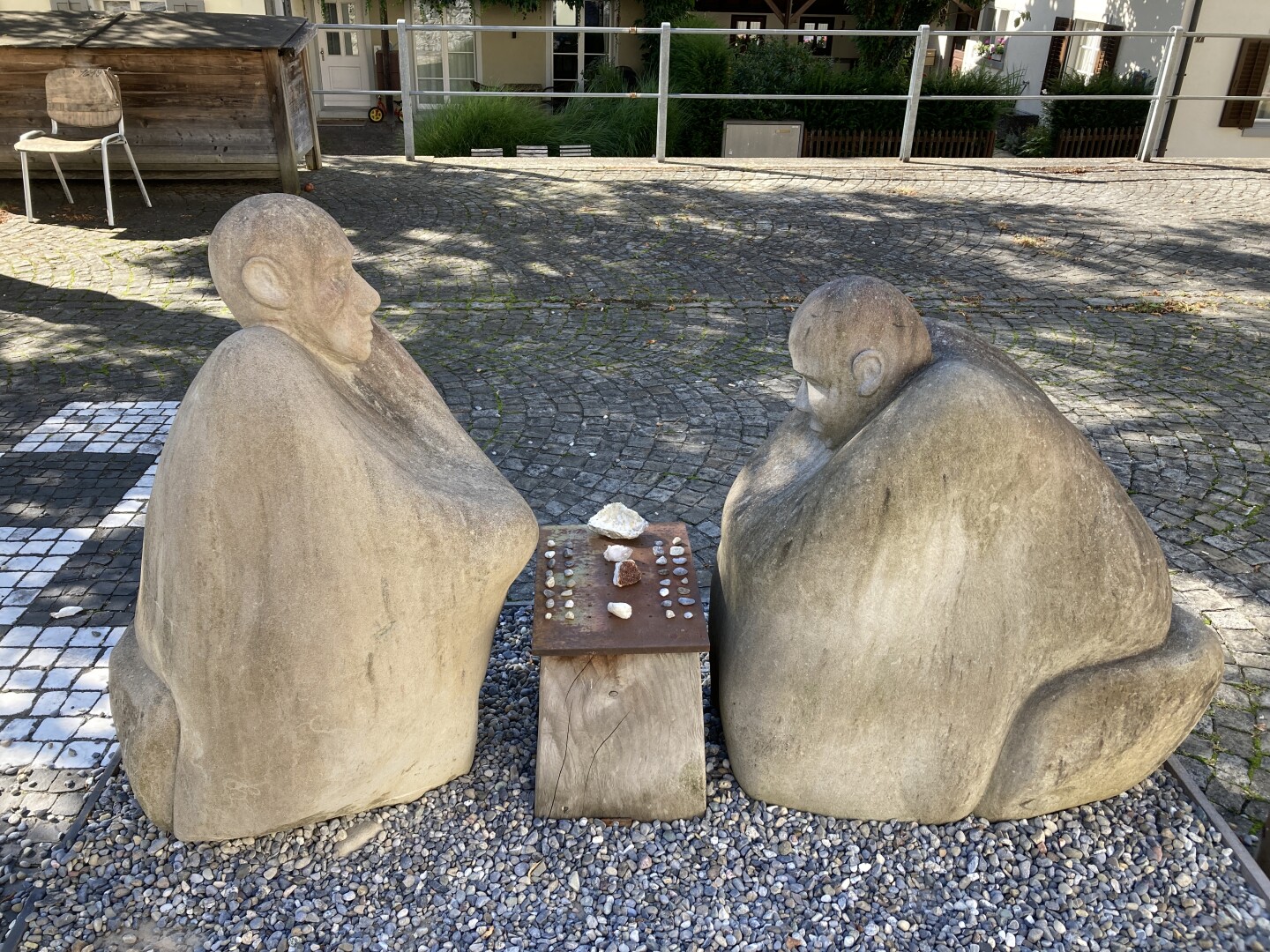 Zwei abstrakte sitzende Stein-Figuren, die an einem kleinen Holztisch ein Brettspiel mit Spielsteinen spielen. Sie befinden sich auf kiesigem Untergrund. Im Hintergrund gibt es Gebäude und Grün, Schatten und Licht, es ist eine sommerliche Atmosphäre.