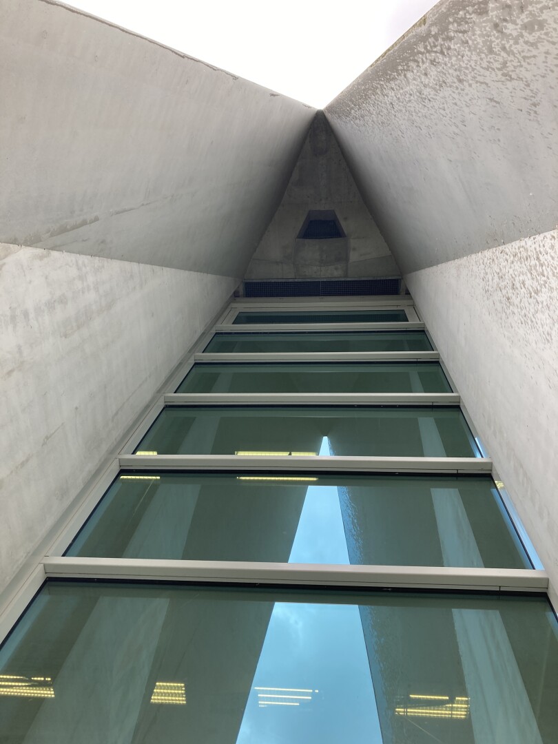 Ein Foto, das einen Teil der Fassade eines hohen, modernen Gebäudes zeigt, aufgenommen vom Boden aus mit Blick nach oben. Betonwände verjüngen sich in Richtung einer spitzen Decke, mit großen Glasfenstern, die den Himmel reflektieren. Das gesamte architektonische Design vermittelt eine schlanke, minimalistische Ästhetik.