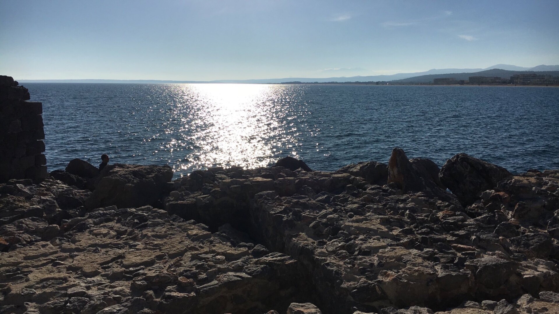 Eine ruhige Küstenszene mit einem felsigen Ufer mit Sonnenlicht, das von ruhigen Gewässern reflektiert wird. Die Kulisse umfasst entfernte Hügel unter klarem Himmel.