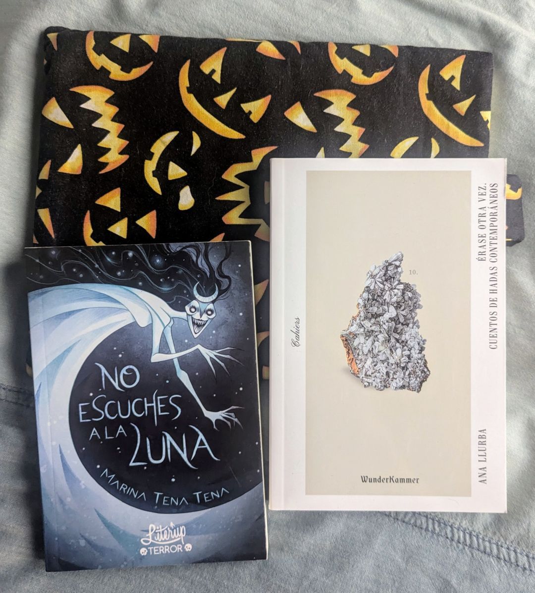 Dos minilibros de editoriales independientes: No escuches a la luna, de Marina Tena Tena (publicado por LiterUp); y Érase otra vez. Cuentos de hadas contemporáneos, de Ana Llubra (publicado por WunderKammer).
Detrás hay una funda de tela halloweenesca de fondo negro y perfiles de calabazas siniestras iluminadas. 
Las portadas de los libritos (que sobresalen muy poco de una mano) se prestan mucho a lecturas de otoño y terror ligero. La de Marina es una especie de luna personificada y siniestra en tonos azulados y oscuros.
La de los cuentos de hadas es sencilla, de tonos claros con una sola imagen encuadrada en el centro que parece una especie de mineral formado por insectos y flores de tonos grises con un punto de color anaranjado en un lateral.