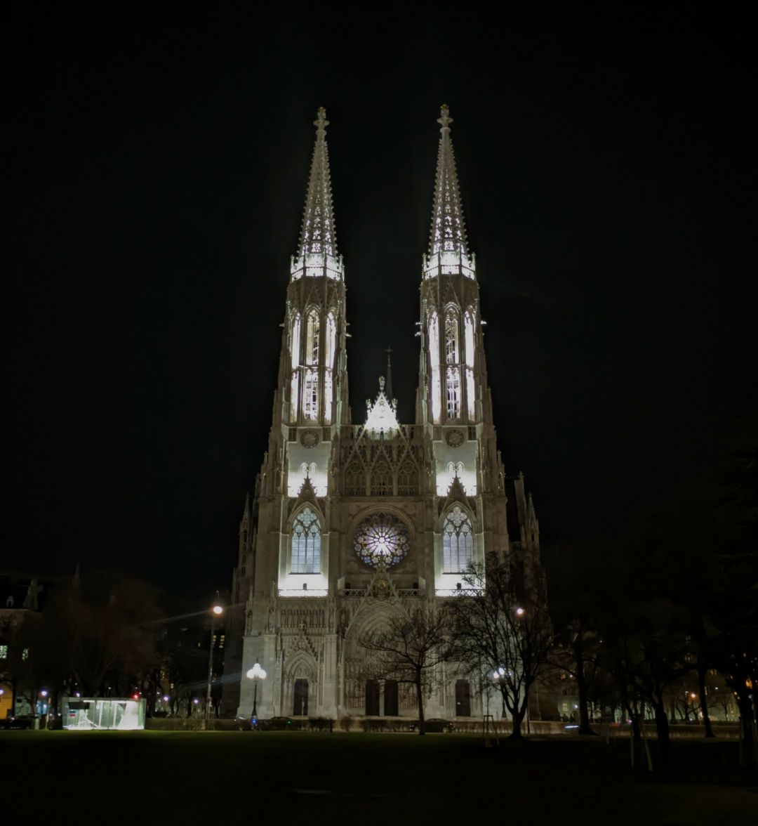 Votivkirche in Vienna - a tall, neo-Gothic, almost white structure with two tall towers in the front. here it is in the dark, with white lights illuminating the front and exaggerating features of the façade