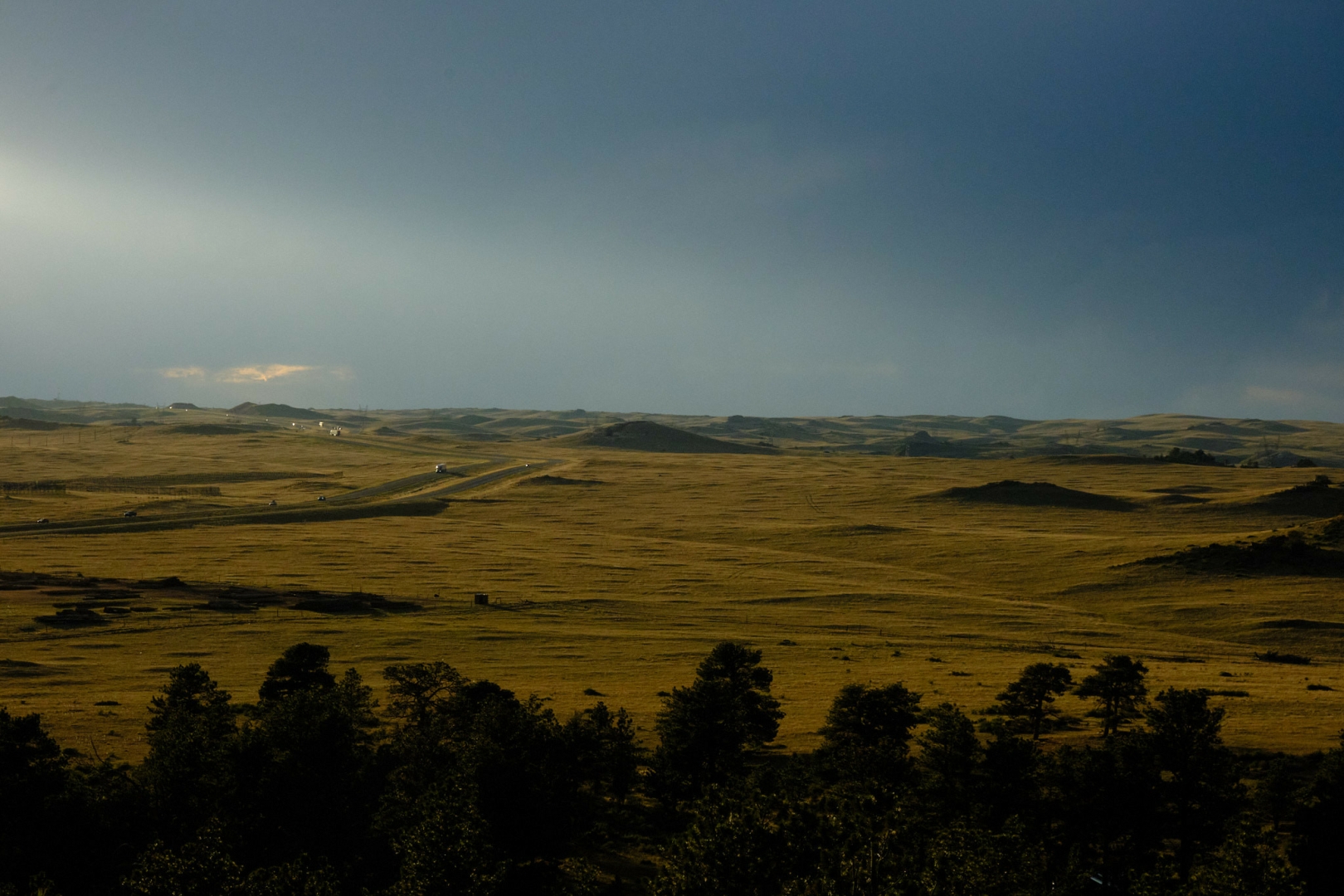 A photo of a barren landscape illuminated by late evening summer light. In the foreground are some trees and to the right of a photo a road winds its way.