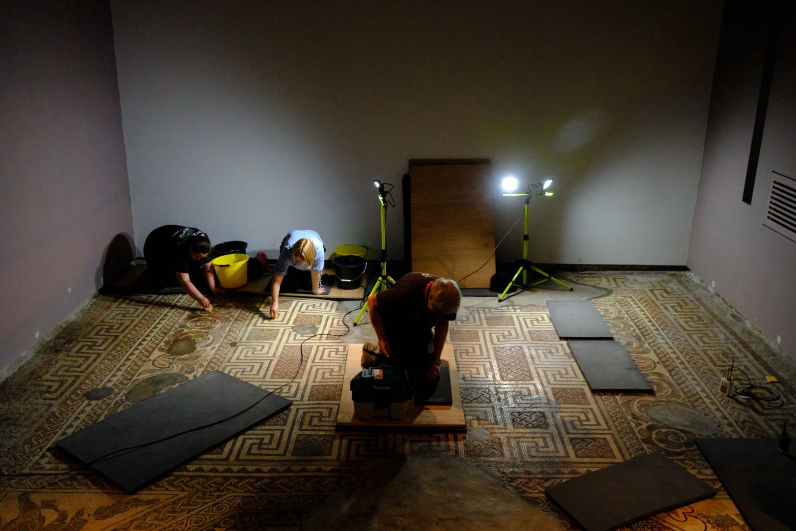 A photograph of a room with a Roman mosaic floor. Three people are cleaning the mosaic using kneeling pads to prevent them from directly touching it. The room is mostly dark but lit by bright spotlights. The mosaic is a mixture of floral and geometric patterns, including swastikas (until the 20th century a symbol of good luck).