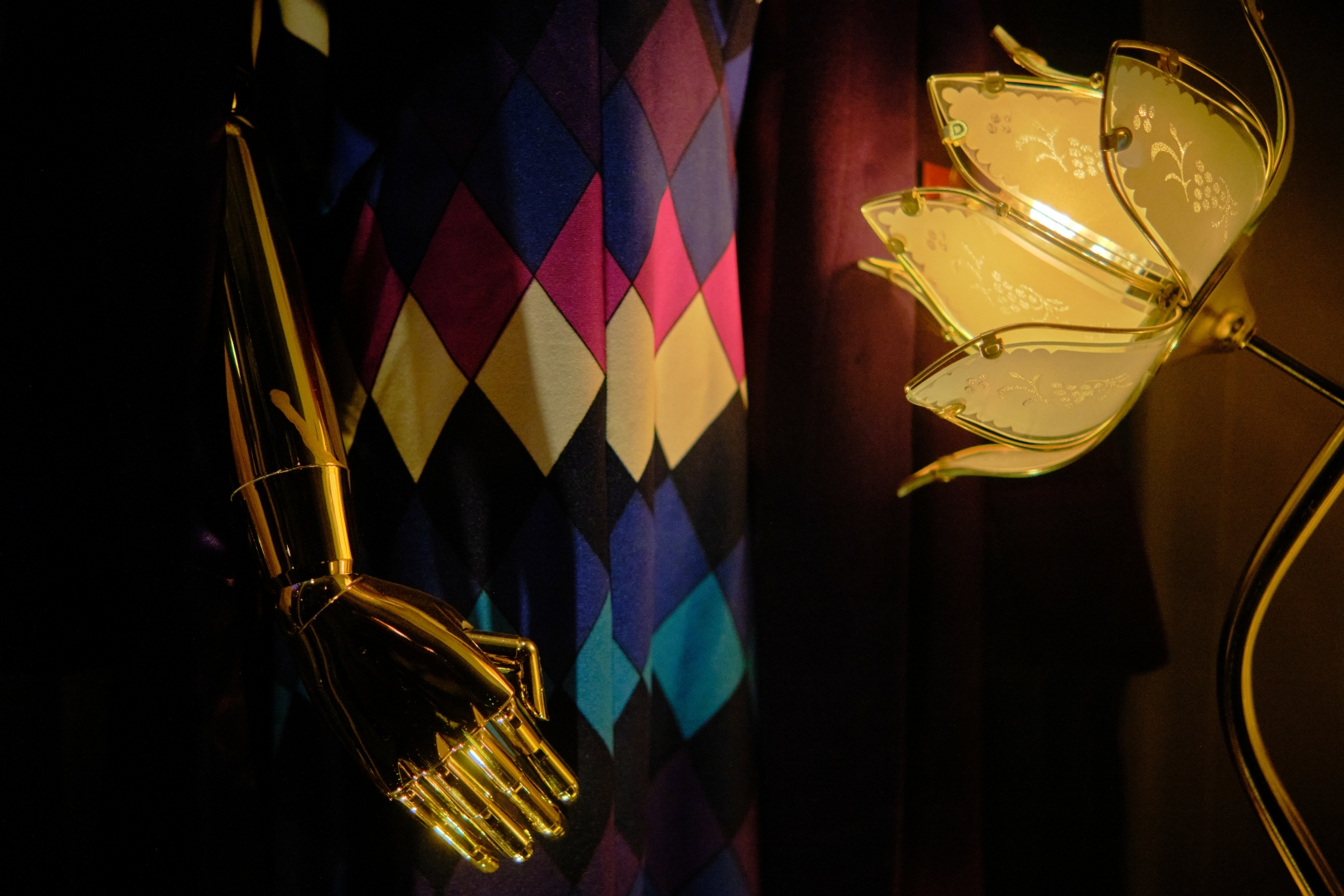 A photograph of a vintage shop window at night. The shot is tightly cropped to take out much of the context leaving a floral glass lamp, a diamond patterned dress and the C3P0-esque hand and arm of the metallic mannequin wearing the dress.