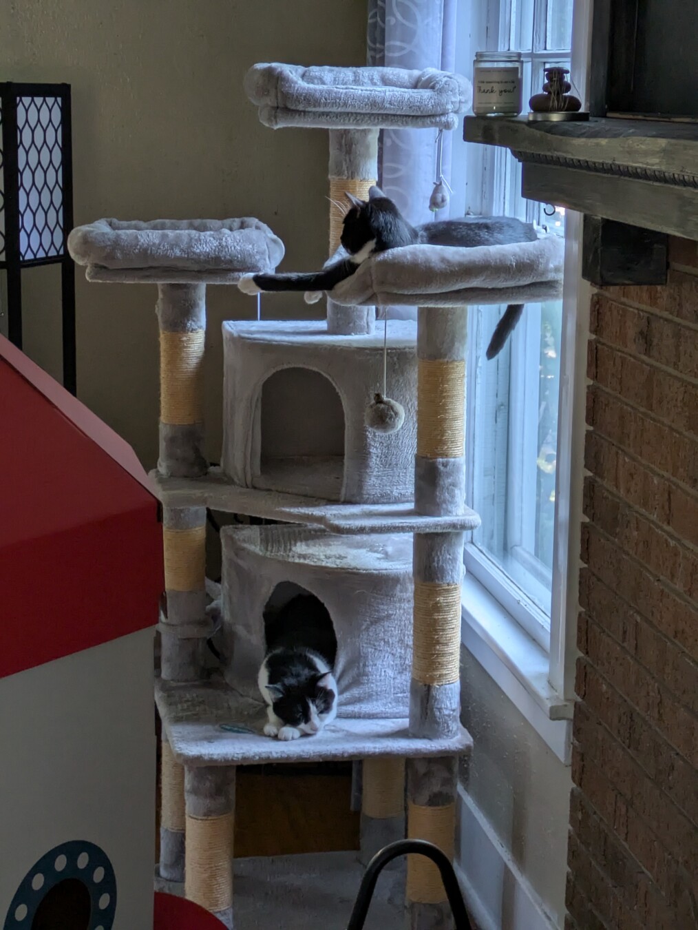 a side view of a large cat tree with two nests. the fabric is gray and the scratching post are covered in yellow twine. it is a little over 6 ft tall. 2 tuxedo cats lie lazily on different parts of the tree.