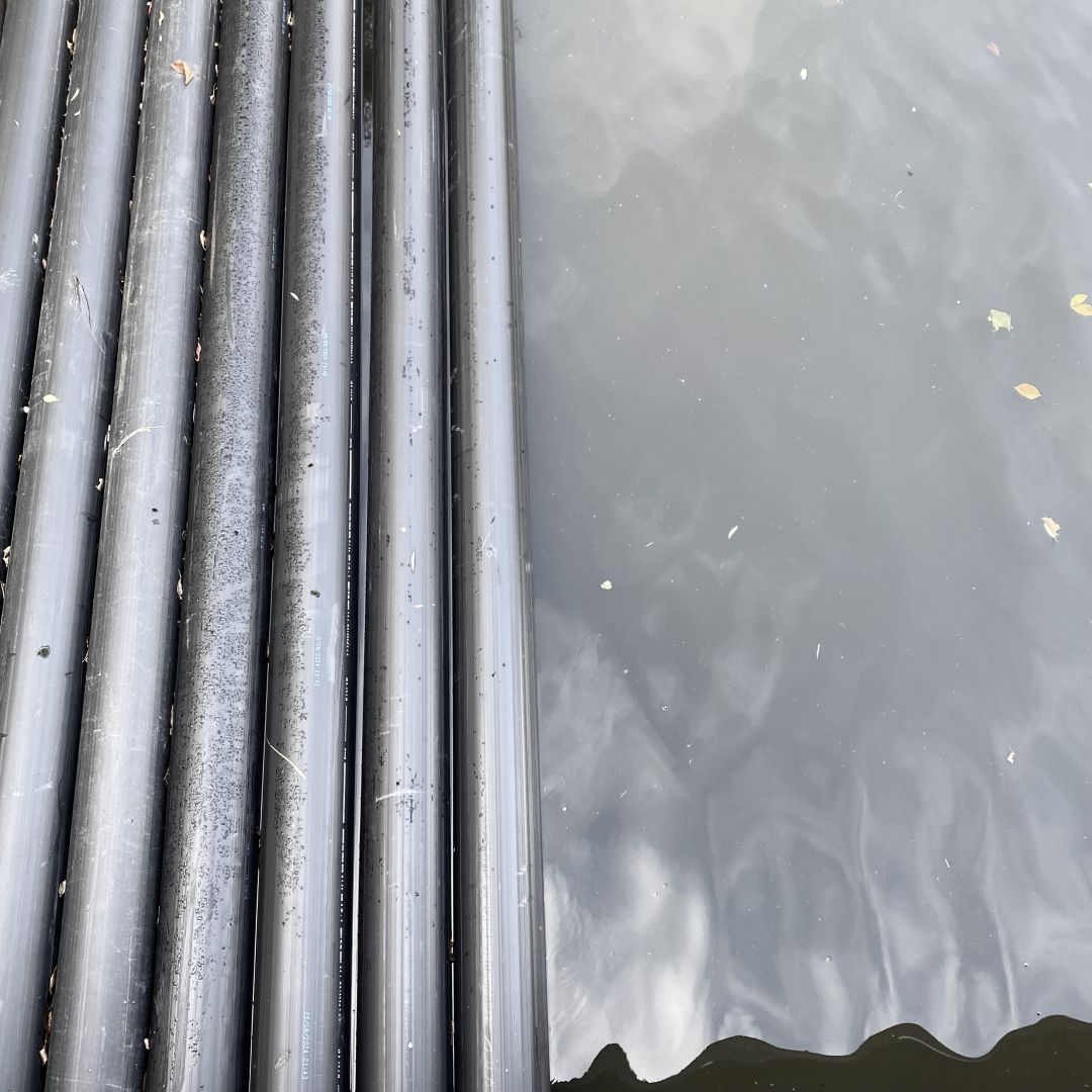 On the left dark grey tubes are running vertically through the photo, floating in a canal. On the right half there is calm water reflecting a grey sky.