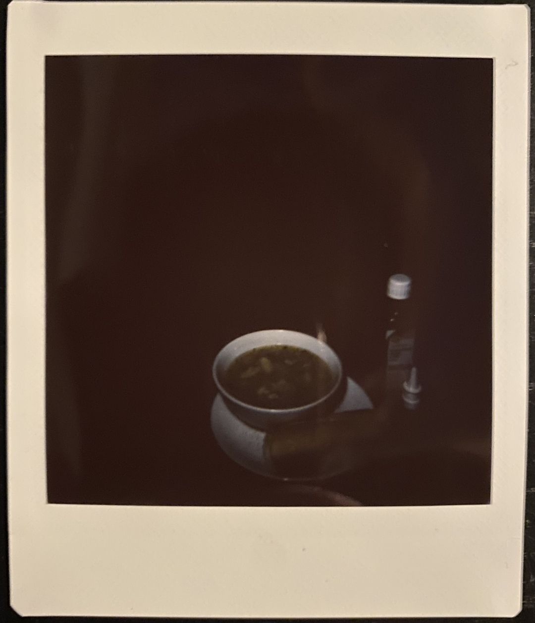 A very dark instant photo with a bowl of soup and non-descript white capped bottle