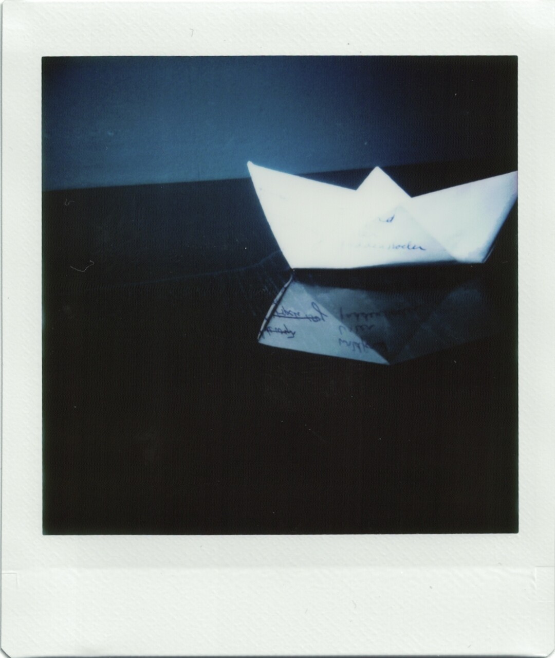 A blown out white paper origami boat in an artificial puddle on a black table. The boat’s reflection shows that it’s made of an old grocery list. 
The boat is a bit to far to the Right of the frame, with its tip just cropped off.