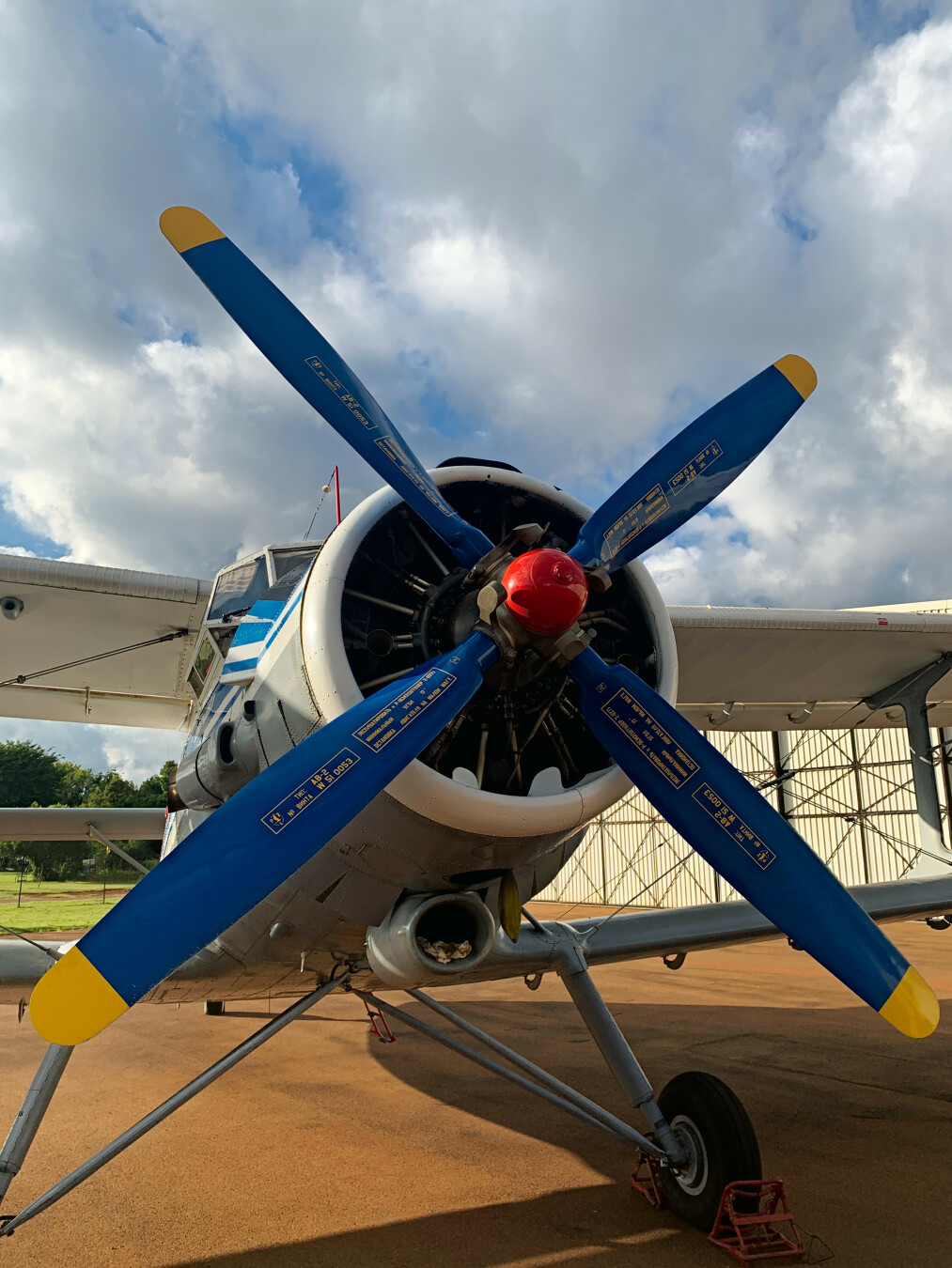 The image captures the imposing front view of an Antononv An-2 airplane, specifically its propeller. The location, determined to be near Wonderboom Airport, in Pretoria, South Africa, is marked by a partly cloudy sky. The focus is clearly on the aircraft, with a well-maintained body and the prominent propeller taking center stage. The surrounding environment includes a glimpse of a hangar and some greenery, indicating an airfield setting. The details on the propeller are visible.