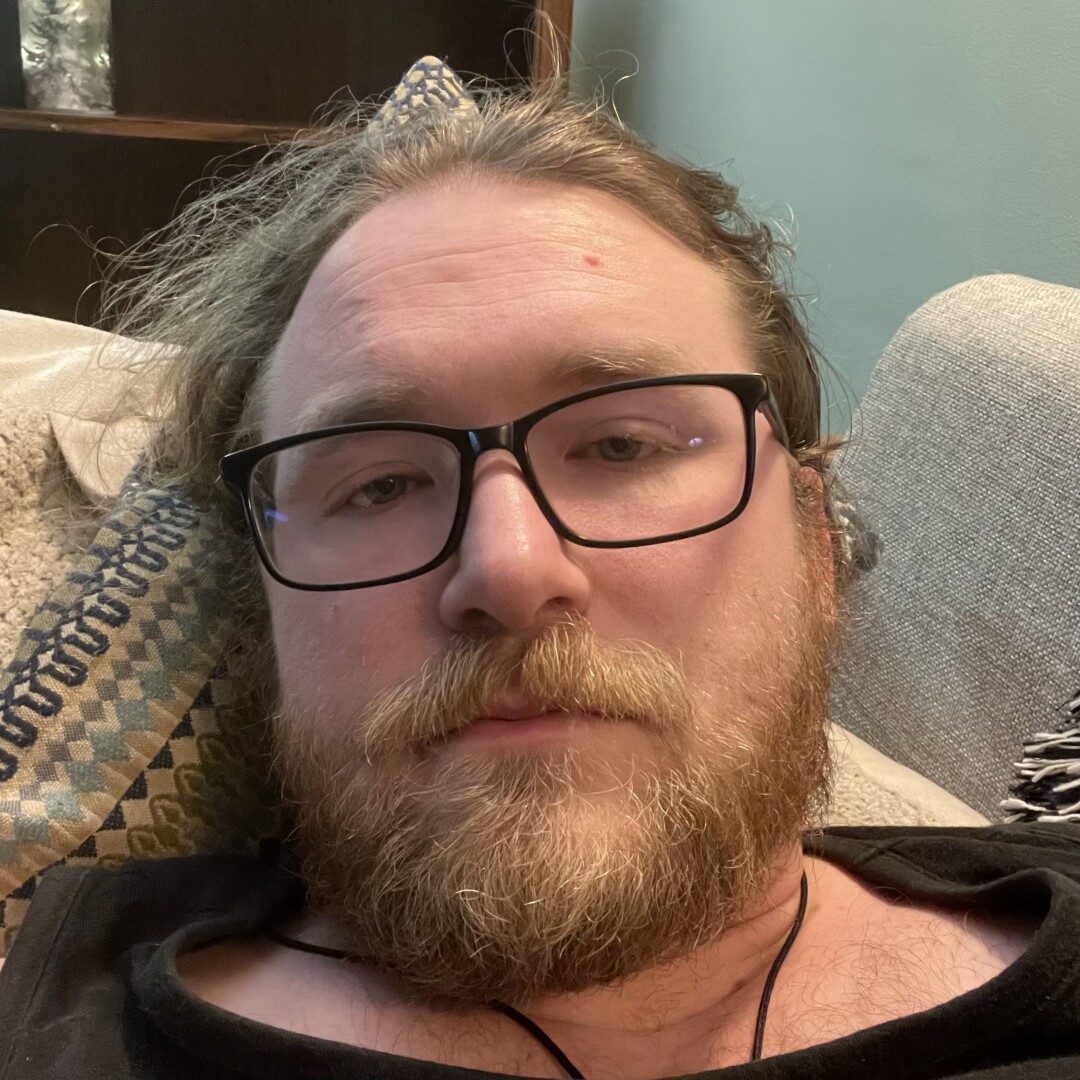 Bearded man with glasses laying on a couch & looking into the camera.