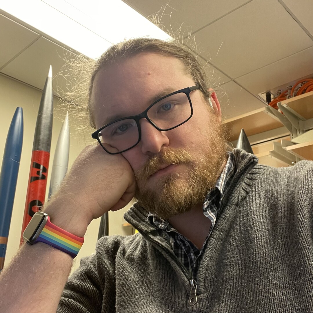 Bearded man in glasses resting his face on his chin & looking into the camera.