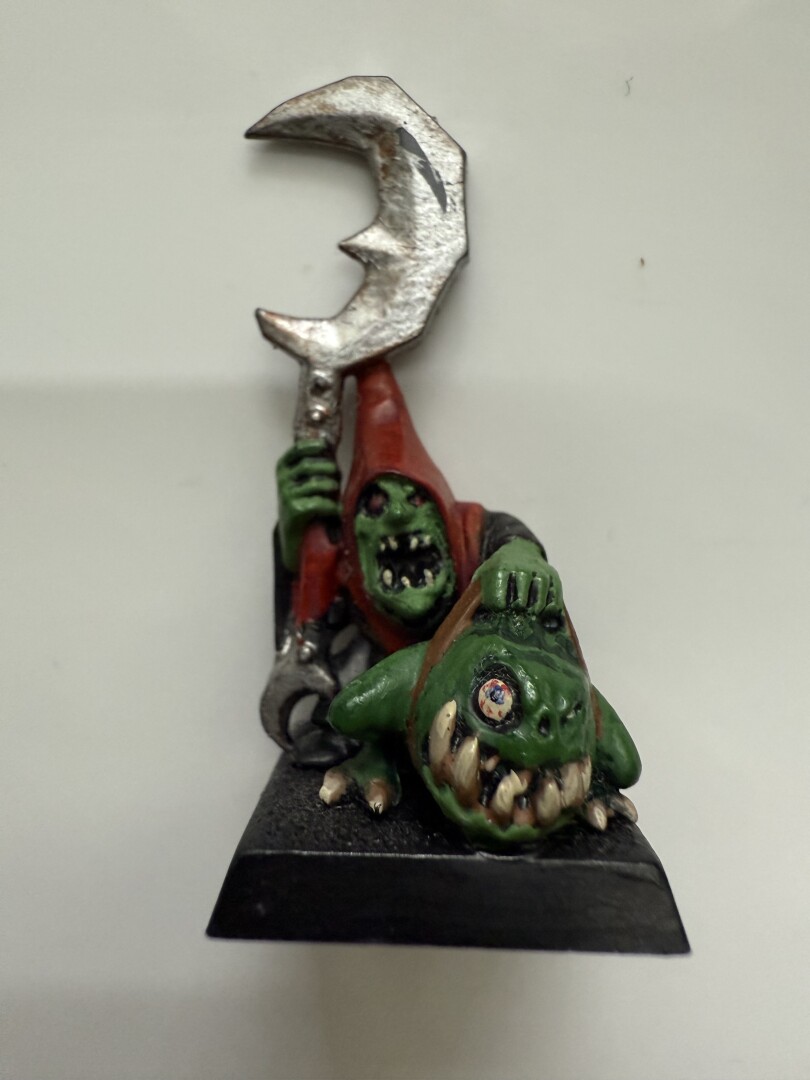 A miniature figure depicts two green-skinned characters; one holds a large, curved weapon and wears a red hooded cloak, while the other is crouched down with an exaggerated expression and sharp teeth.